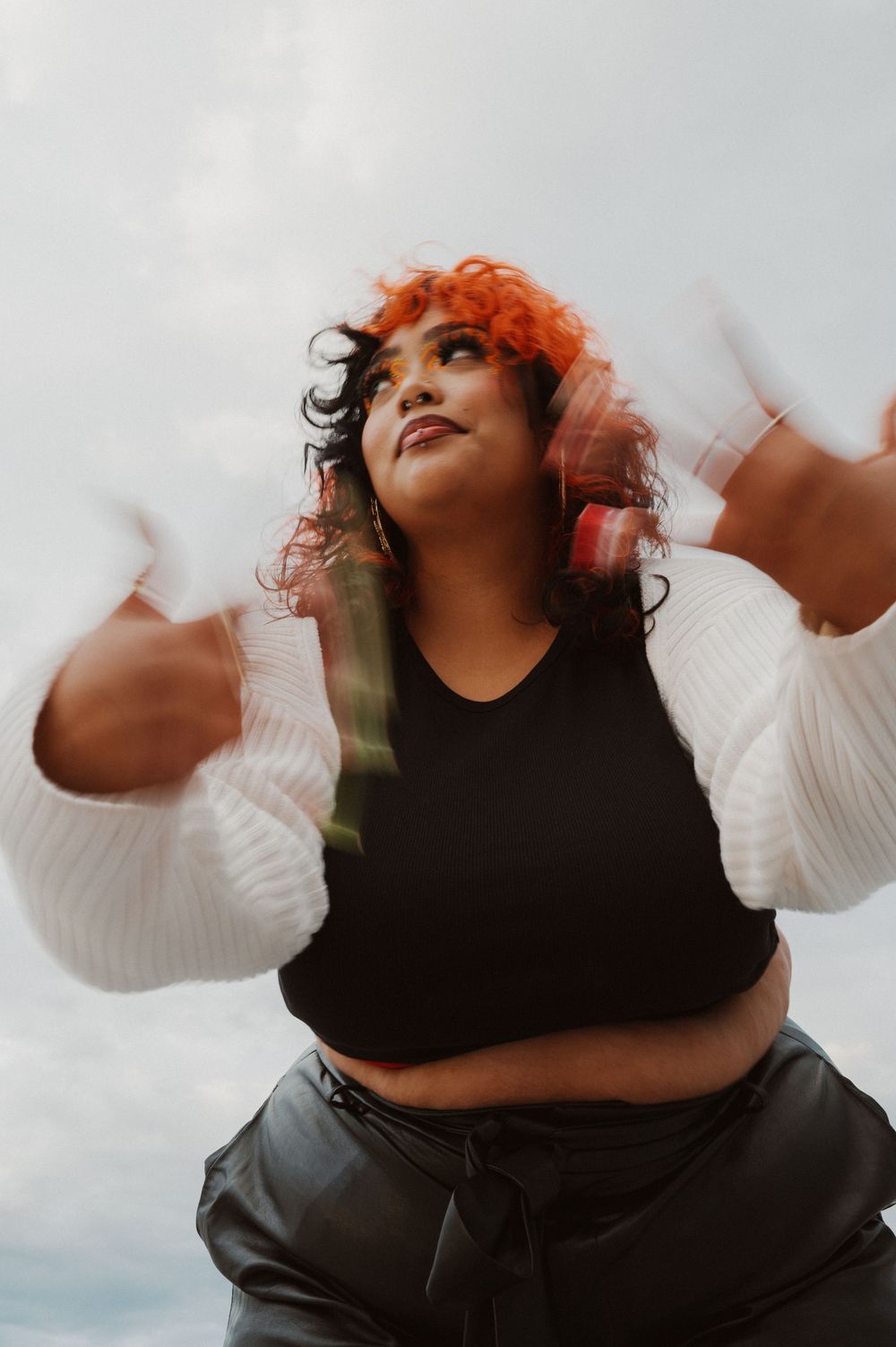 Low angle photo of person with bright orange curly hair wearing a cropped black top and pants against cloudy sky.