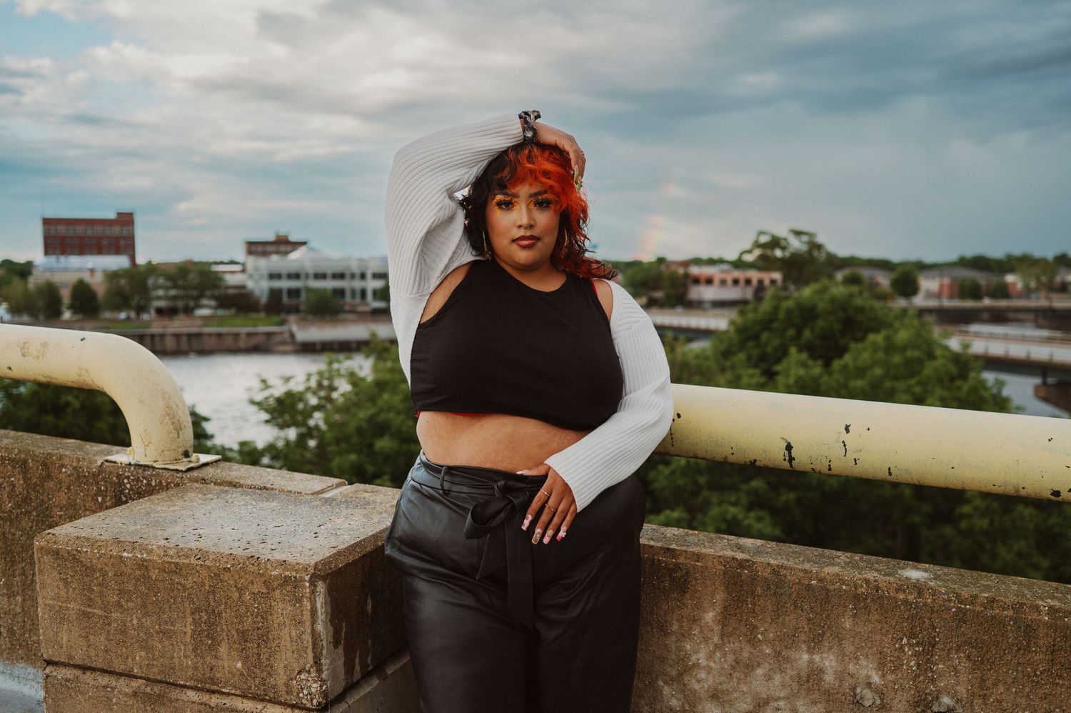 A woman in a black dress poses confidently on a concrete rooftop with an urban skyline in the background.