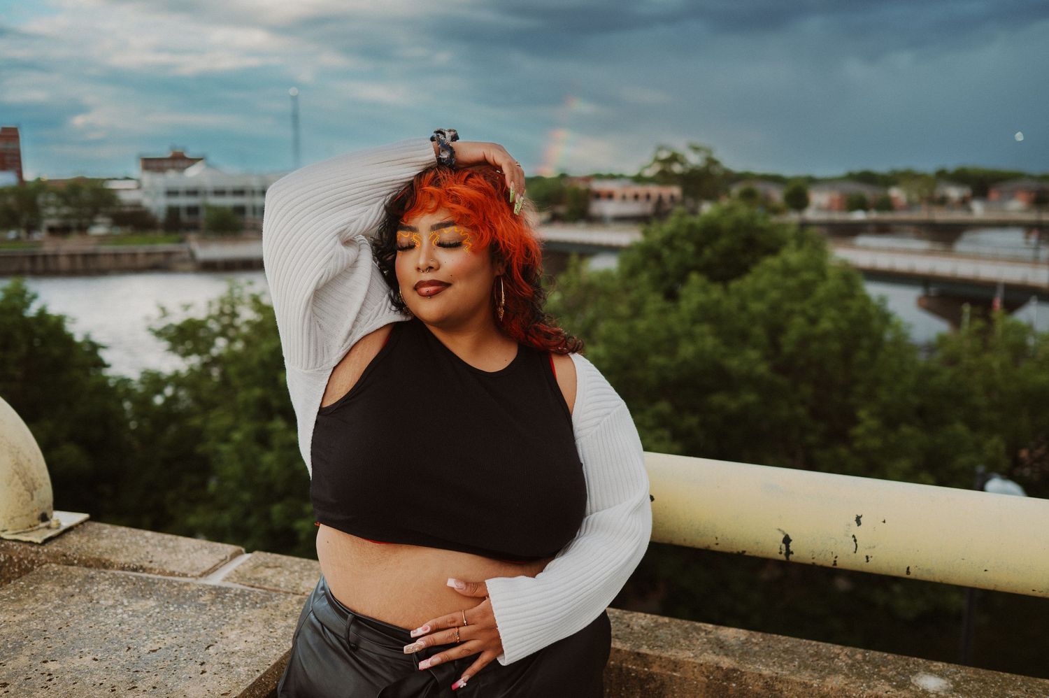 Person with vibrant red hair poses outdoors on a rooftop wearing a black crop top with white sleeves against a cloudy sky.