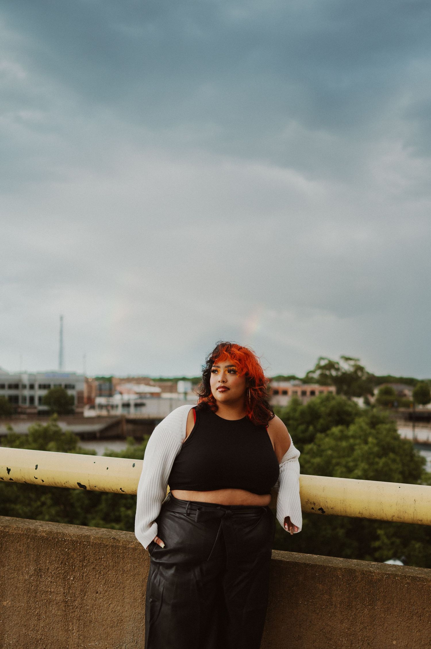 A plus-size person in black clothing poses confidently on a concrete wall against a cloudy urban skyline background.