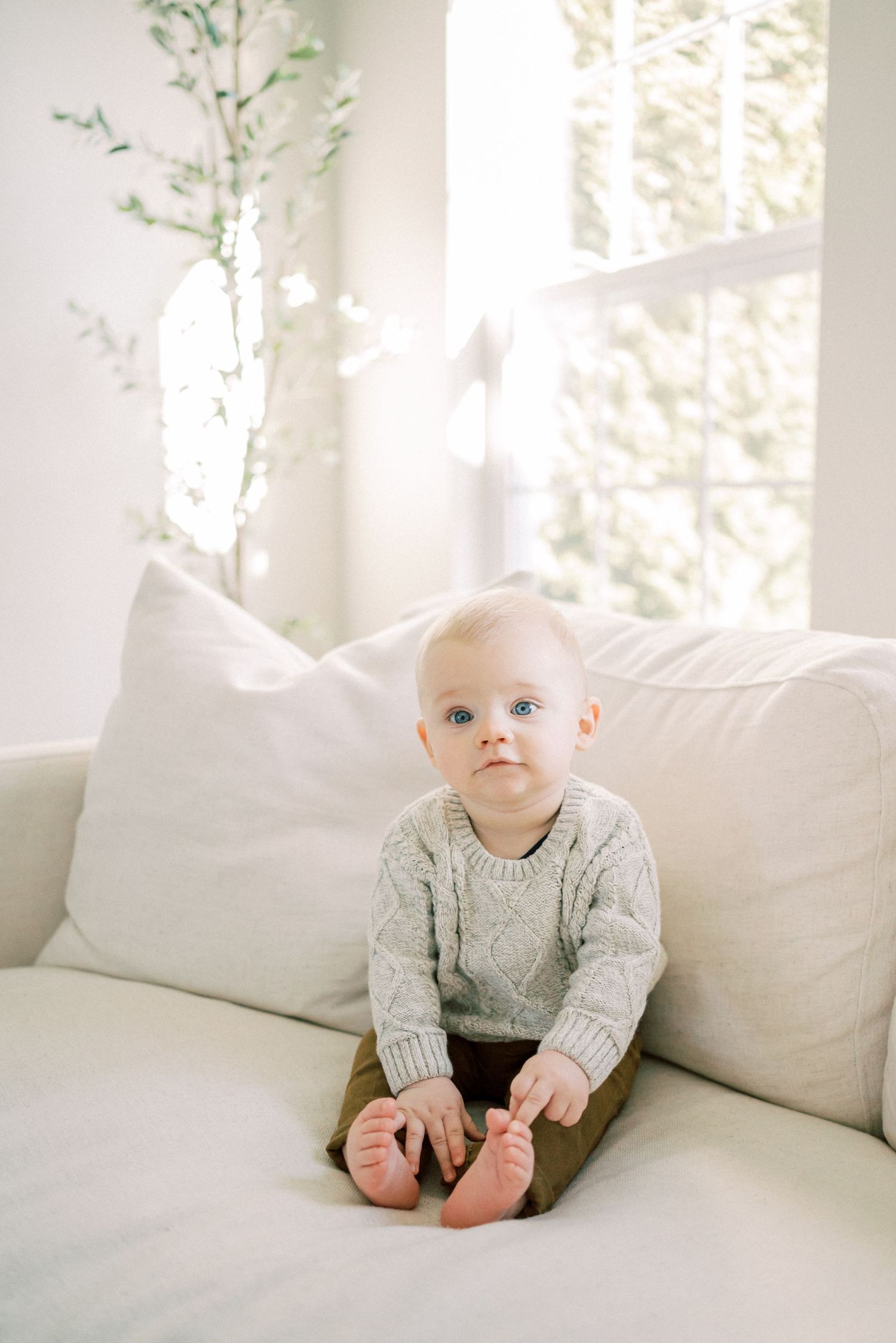 Baby wearing knit sweater sits contentedly on white couch by bright window.