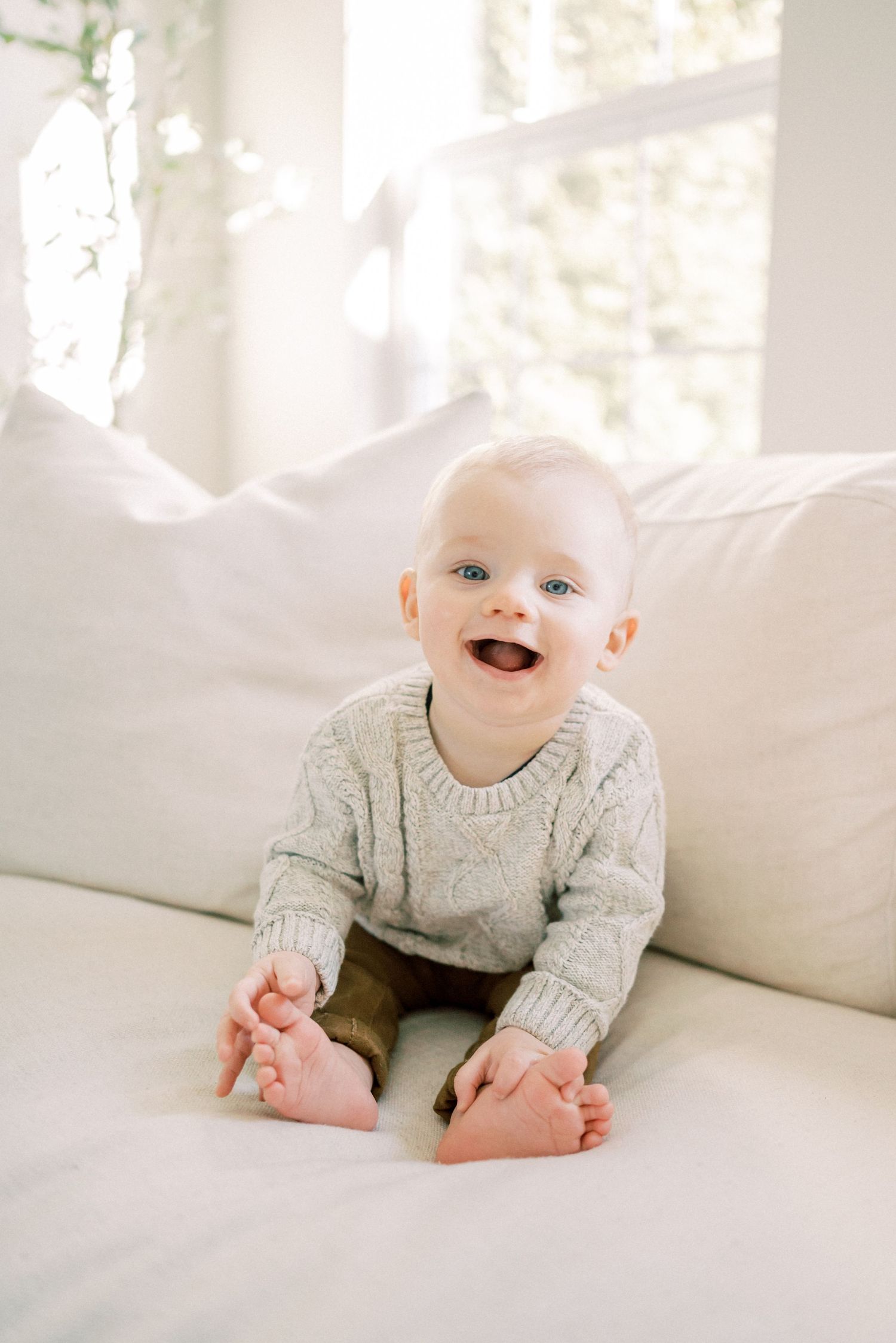 Happy baby in gray sweater sitting on white couch near sunny window.