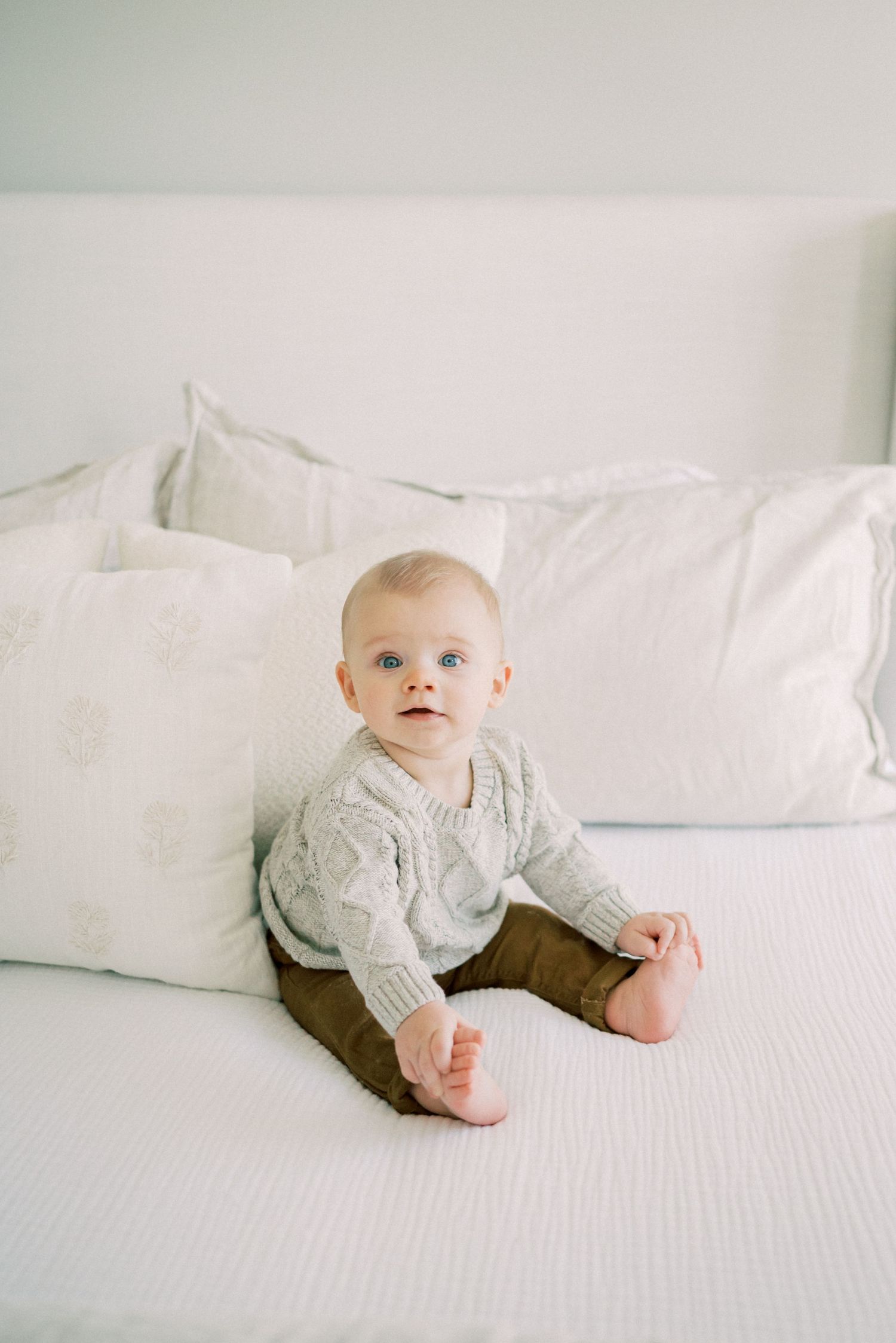 Baby in grey sweater and brown pants sits on white bed with pillows.
