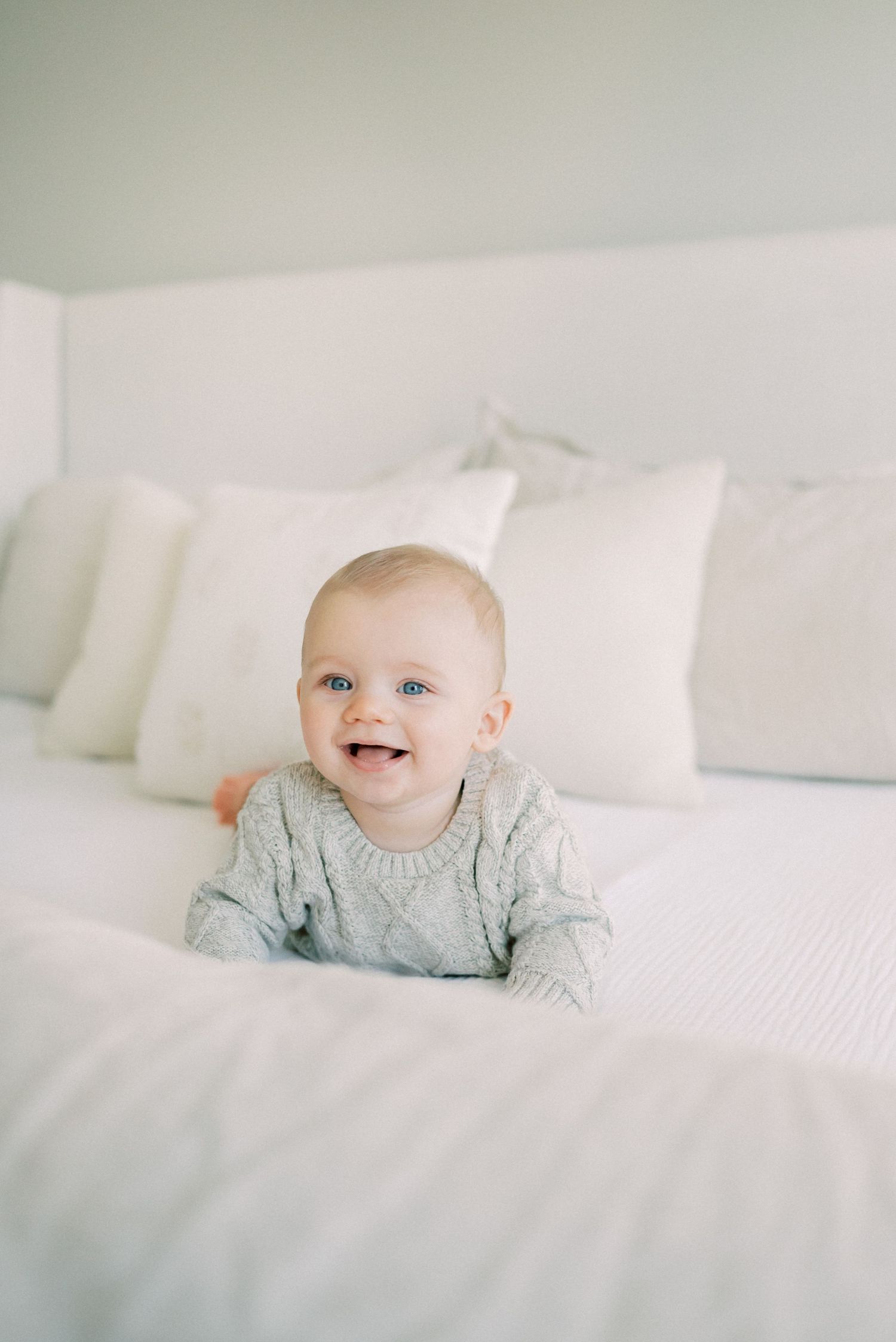 Smiling baby in grey sweater lying on white bedding.
