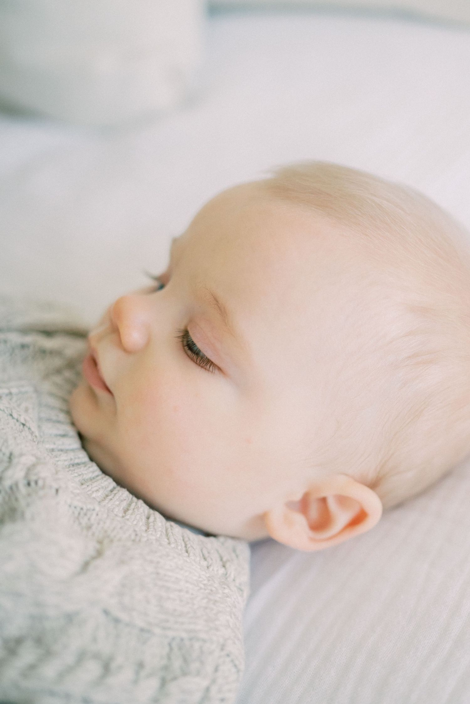 Sleeping baby in grey knit sweater resting on white bedding.