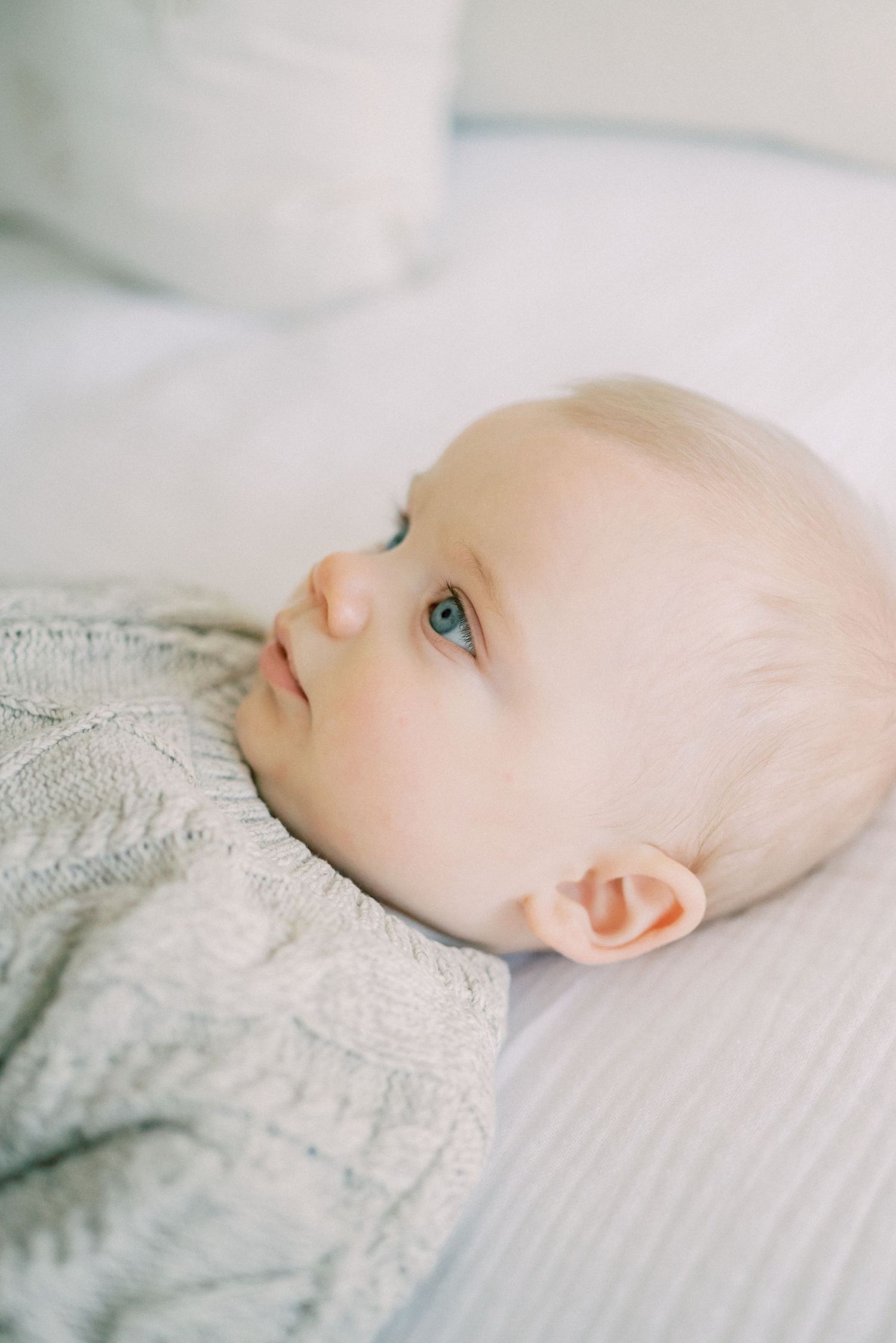 Close up of baby with bright blue eyes wearing grey sweater on white bedding.