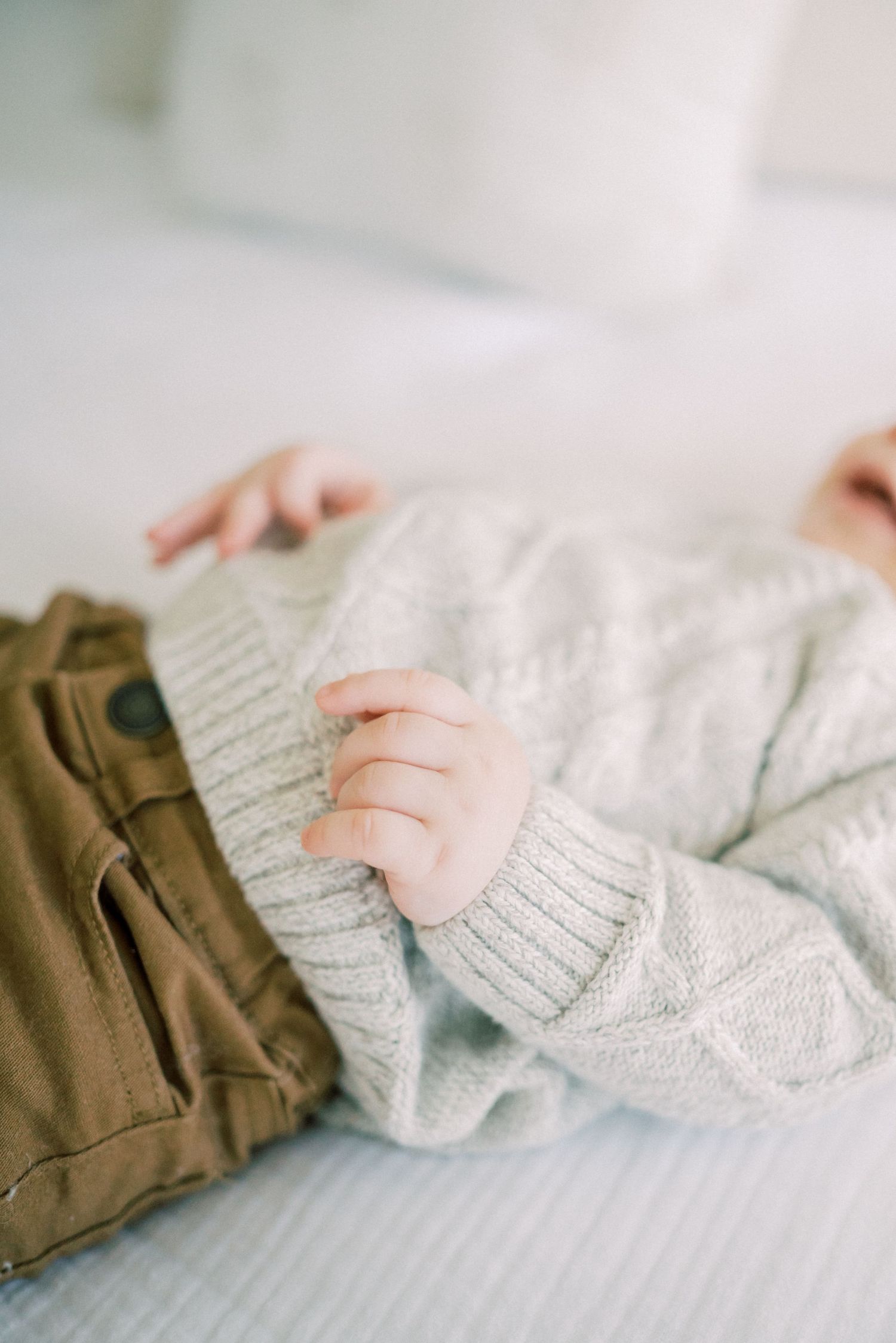 Detail shot of baby wearing grey sweater and brown pants on white bedding.
