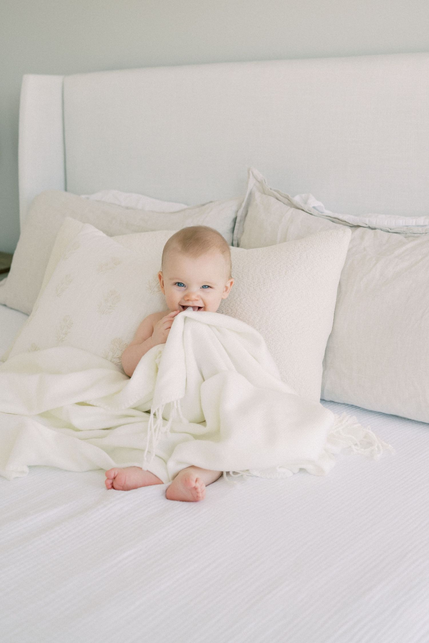 Baby in white playing with blanket on crisp white bedding.
