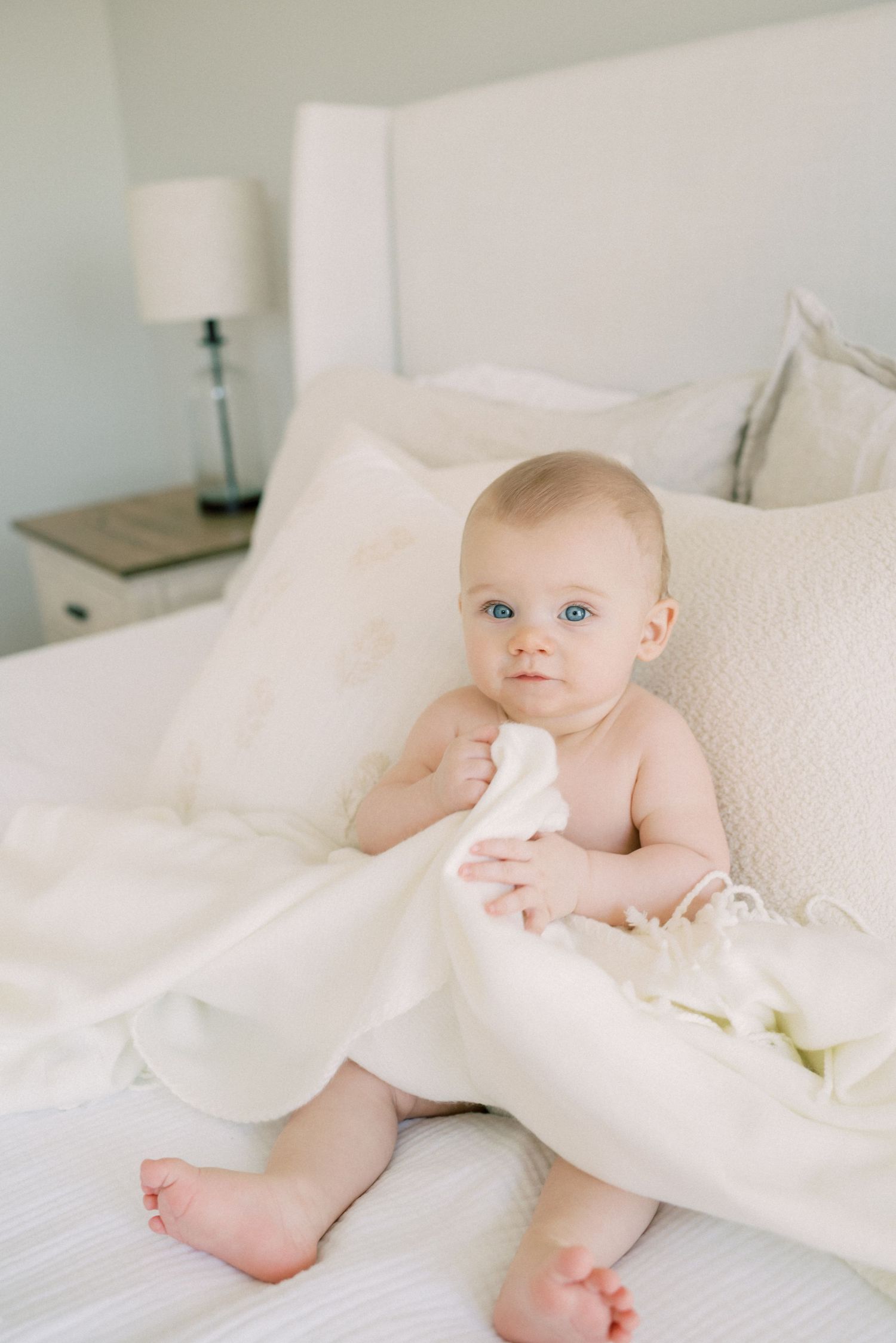 Baby with bright blue eyes sitting among white pillows and bedding.