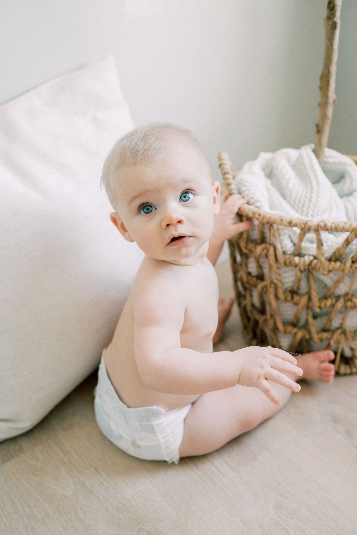 Baby in a diaper sits on floor near white cushions and woven basket.