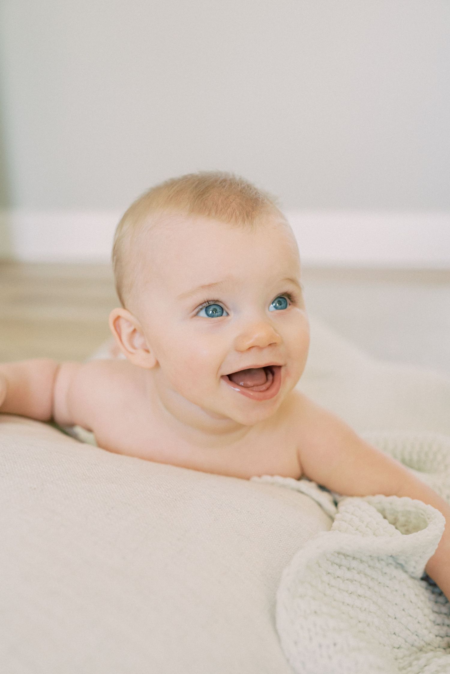 Joyful baby with bright blue eyes and blonde hair smiles while lying on white bedding.