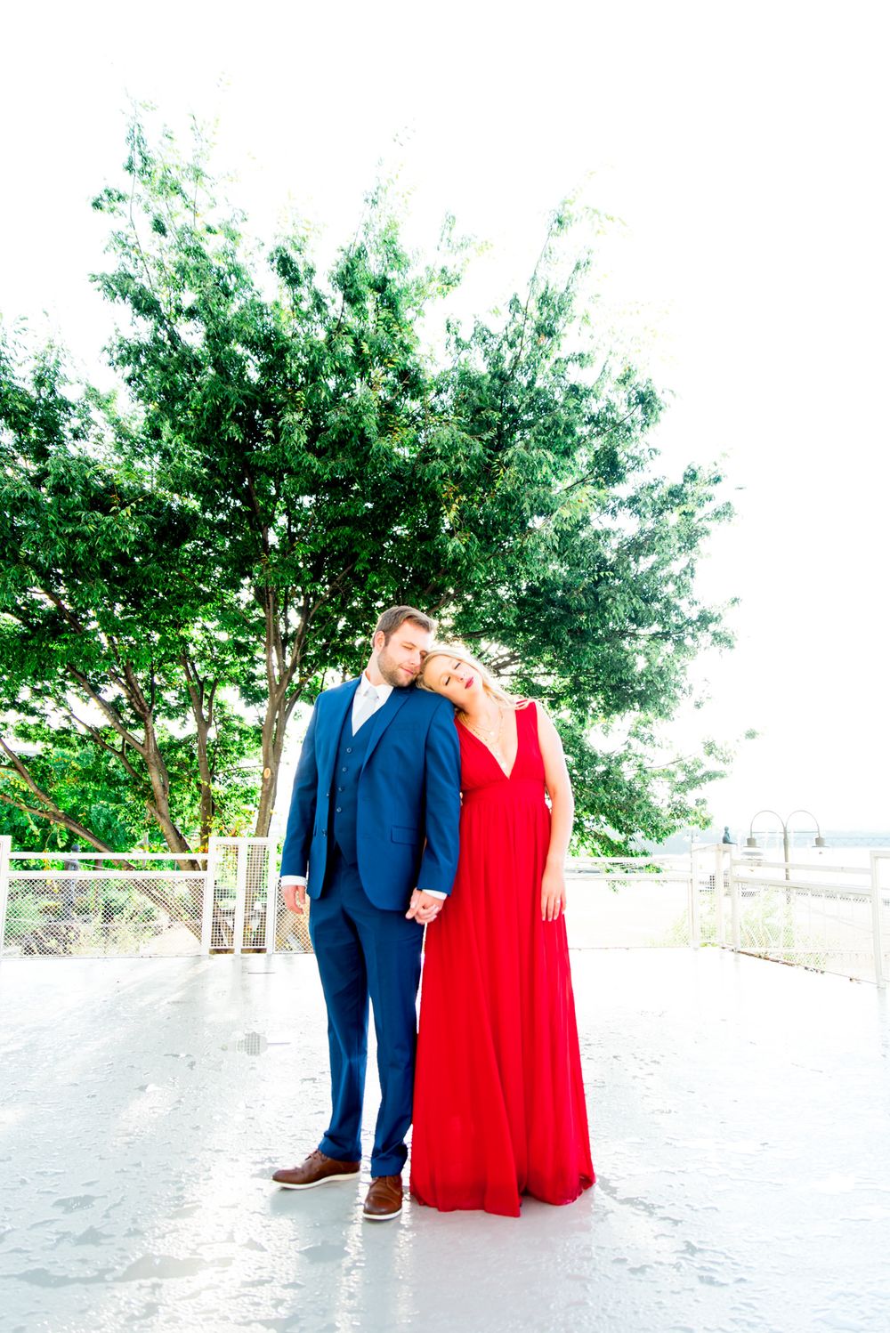 blonde woman in red dress and man in navy suit lean on each other with eyes closed in front of big tree