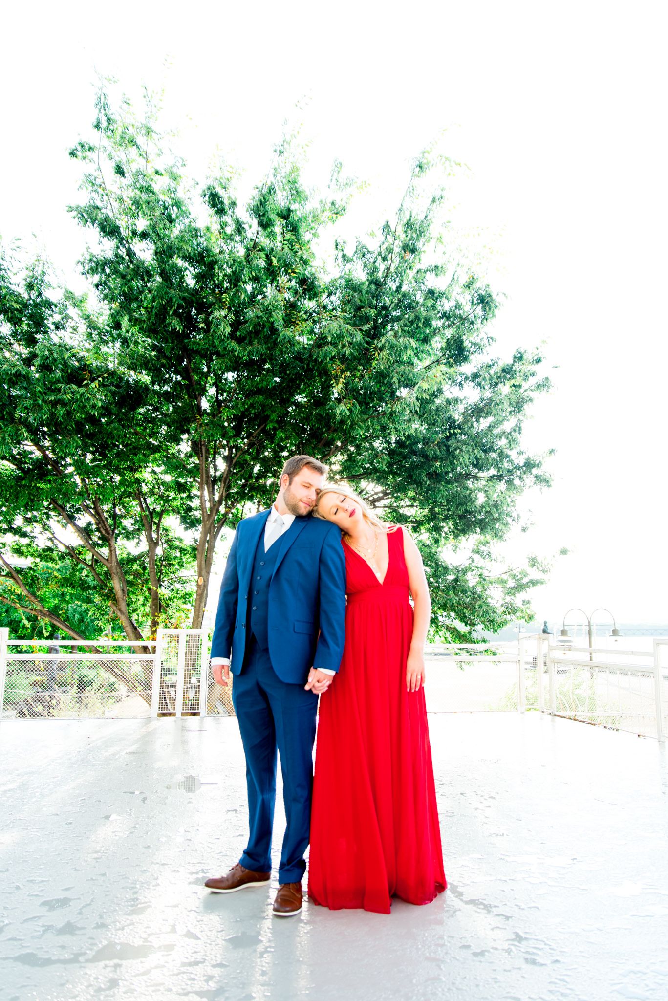 blonde woman in red dress and man in navy suit lean on each other with eyes closed in front of big tree