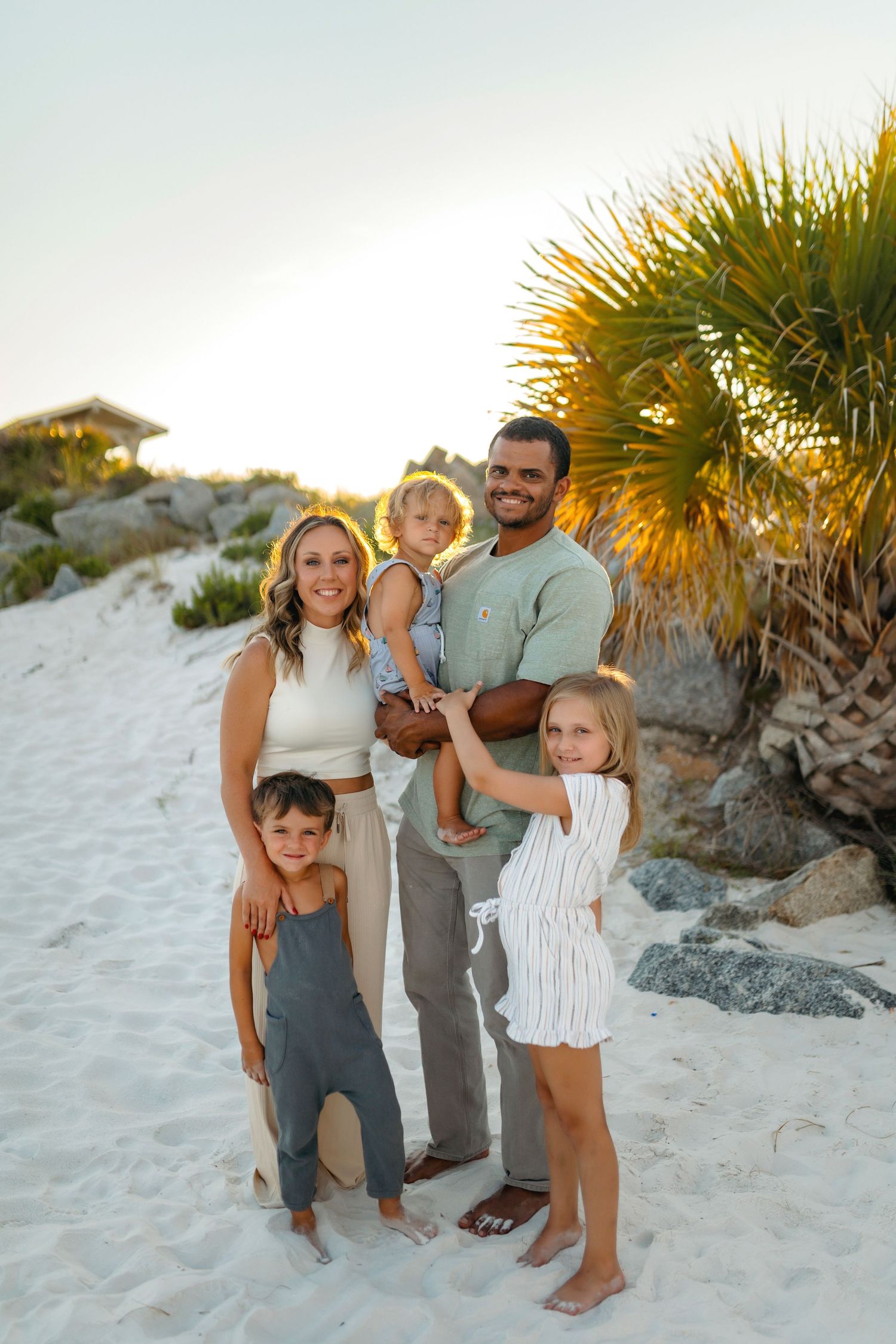A happy family poses together on a sunny beach with palm trees and sand dunes in the background.