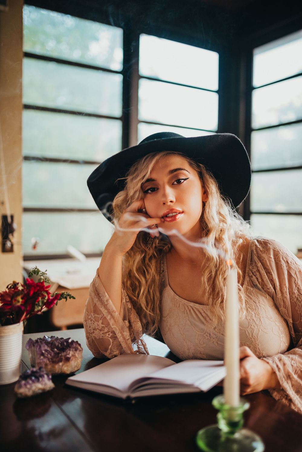 Person in black hat sitting at table with book and candle in sunlit room.
