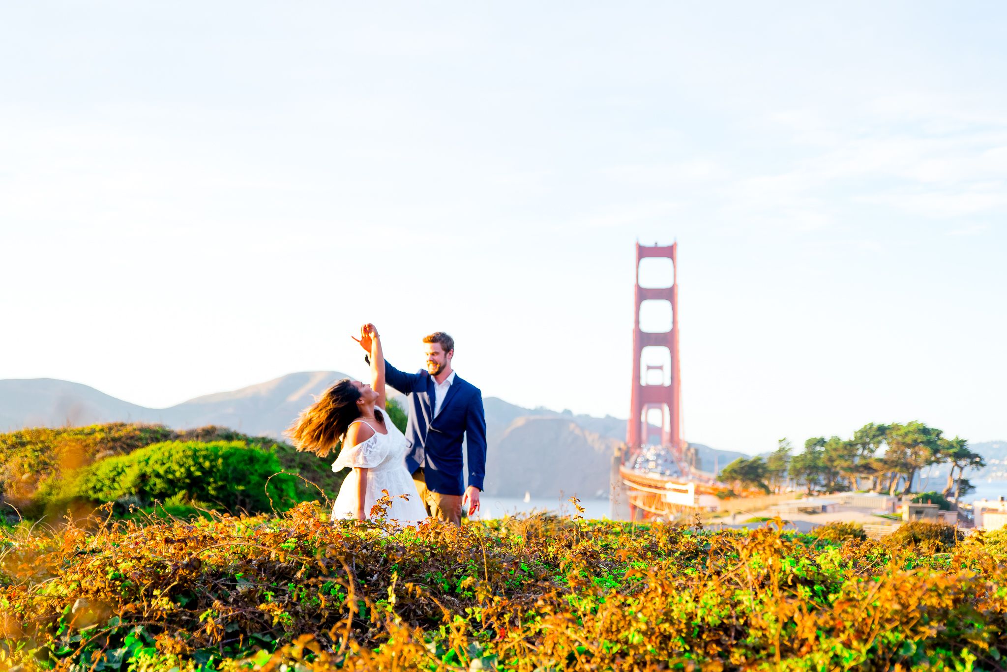 man in navy suit twirling woman in white dress at sunset in hills in front of Golden Gate Bridge in background