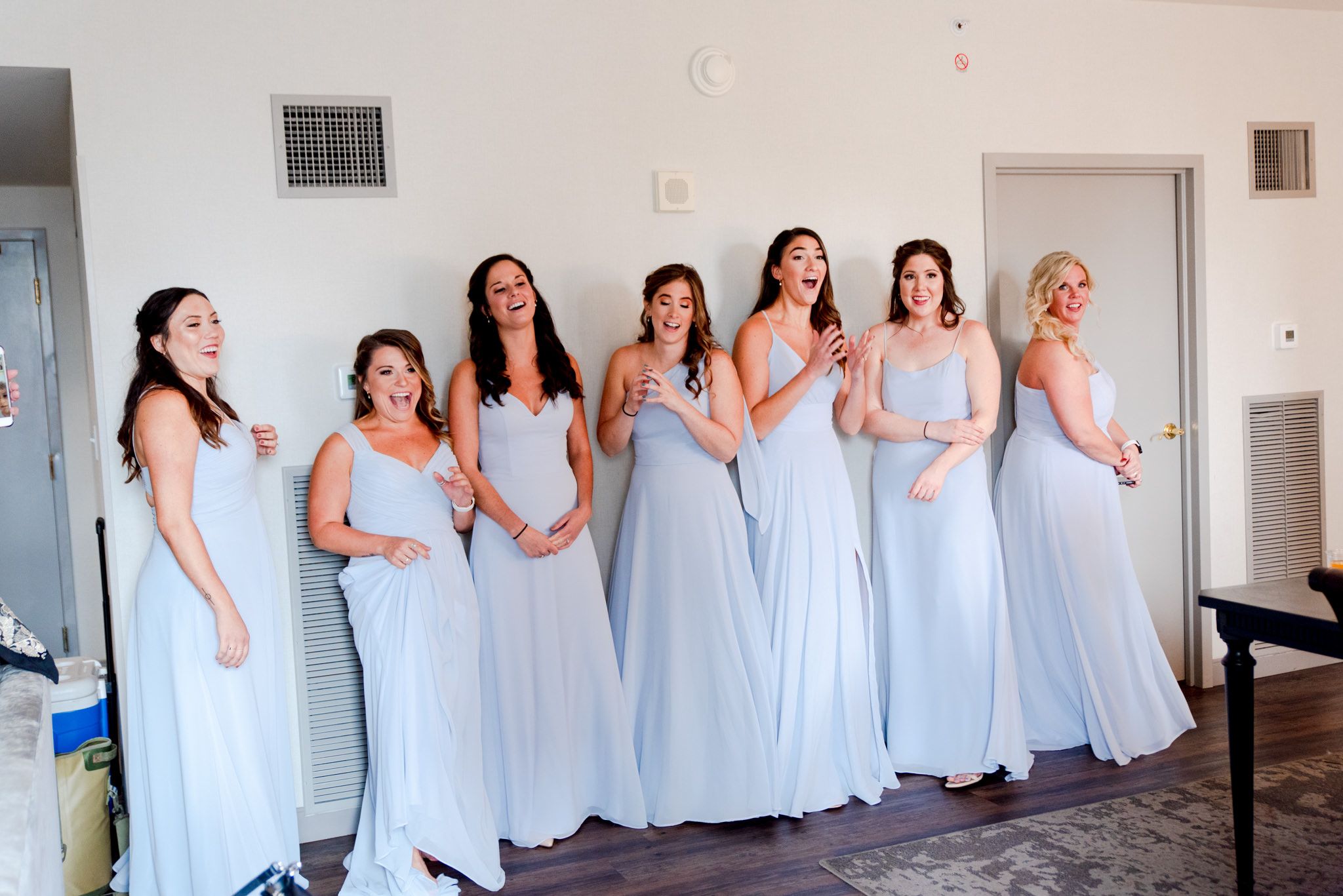 7 bridesmaids in light blue dresses smiling and clapping after seeing the bride, while mom smiles and holds up phone
