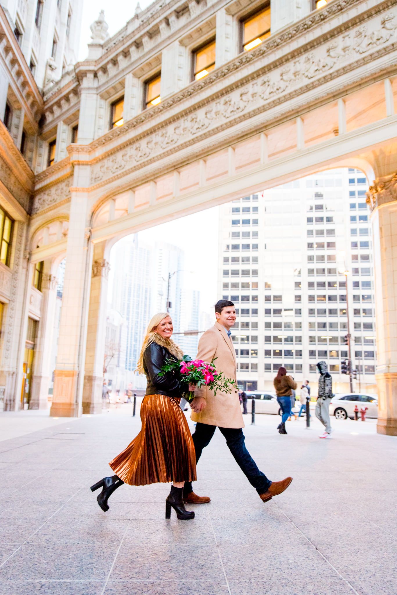 man in tan coat walking with woman in copper skirt in front of Wrigley Building engagement photos