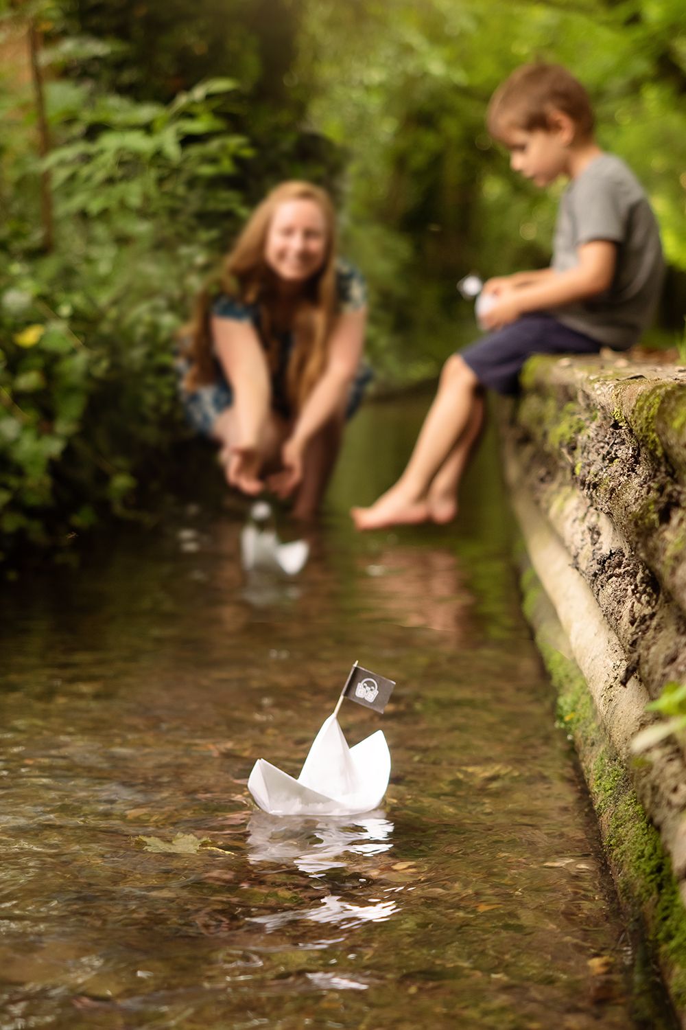 A woman and child sail paper boats along a small stream