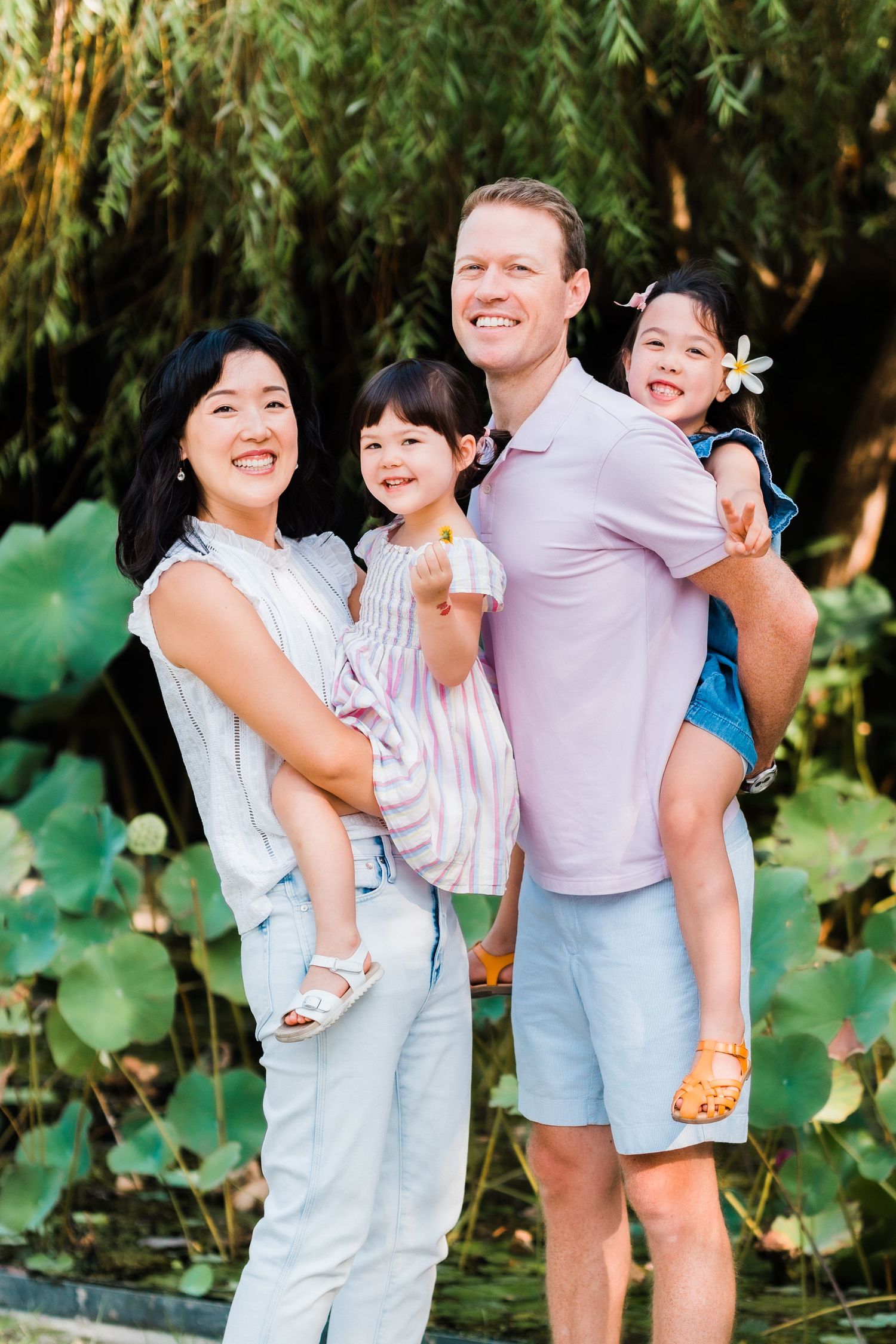 A cheerful multicultural family poses together outdoors against a backdrop of willow trees and lush greenery.