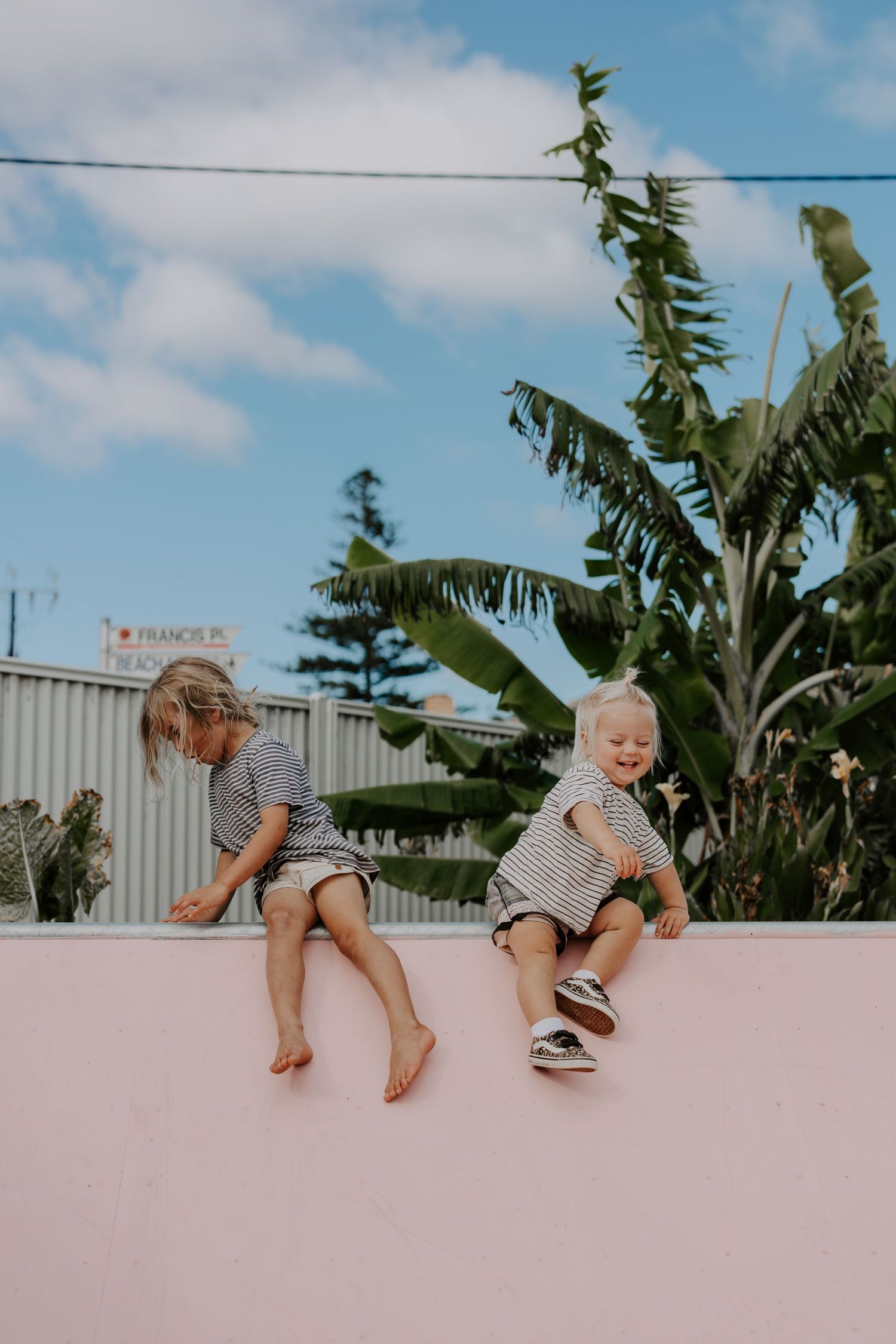 Children sitting on pink wall with palm trees behind them under blue sky.