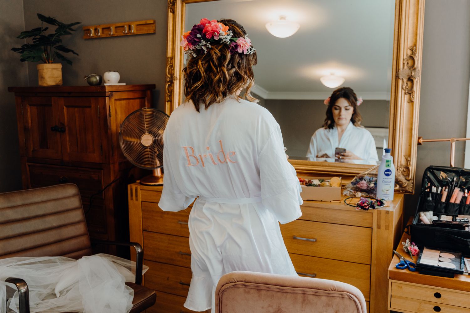 Wedding preparation scene with white robe reflected in vintage wooden vanity mirror.