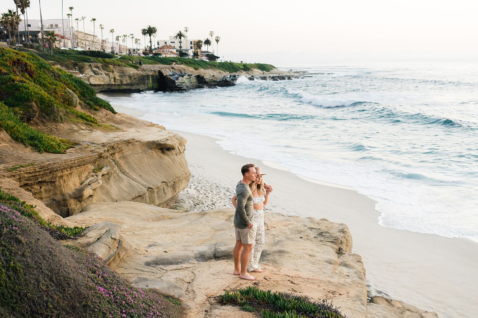 Couple standing on rocky coastal outcrop overlooking Pacific ocean with palm trees in distance.