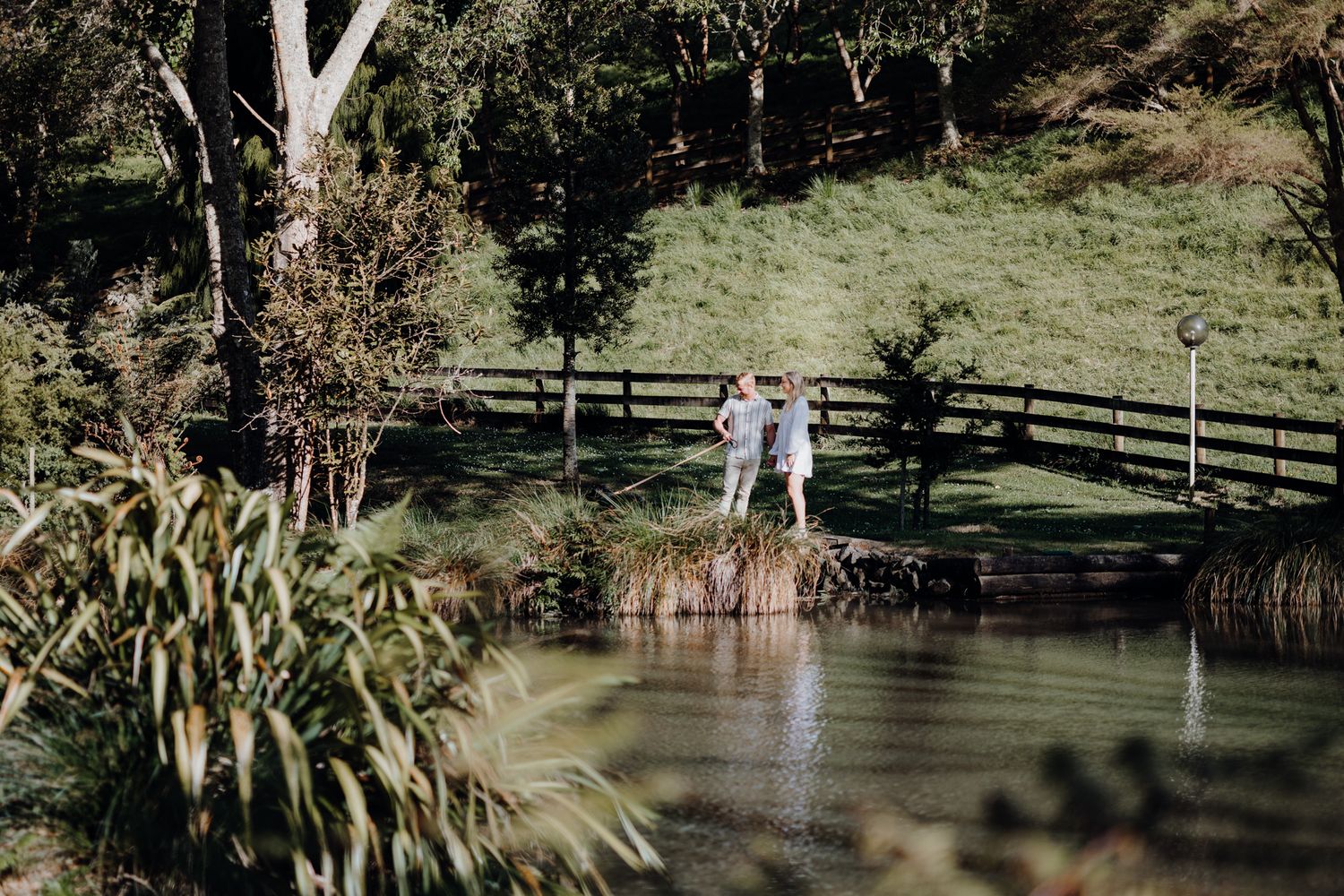 Two people stand together on a wooden dock overlooking a peaceful pond surrounded by nature.
