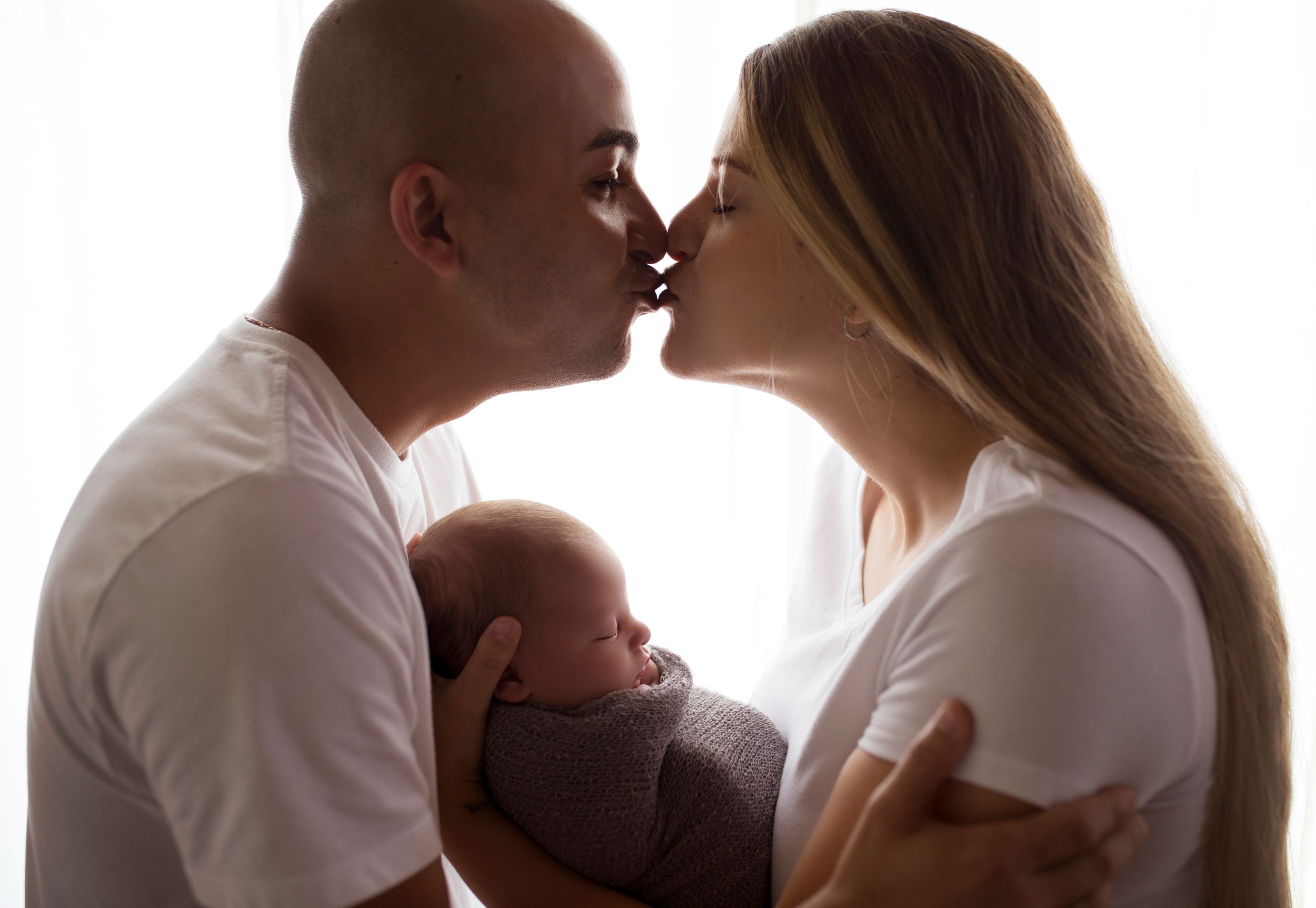 couple holds child while kissing image is backlit