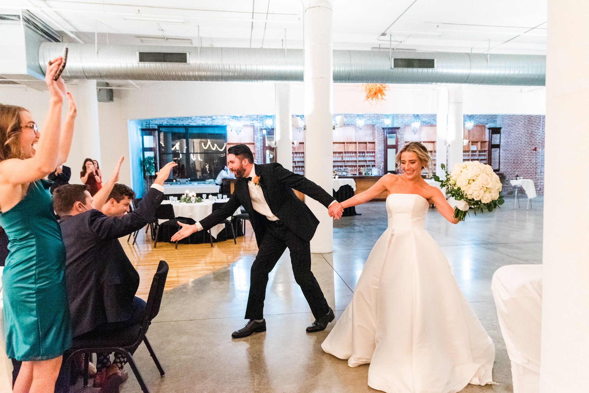 bride and groom walk into wedding reception area smiling, dancing, holding hands, and high fiving people