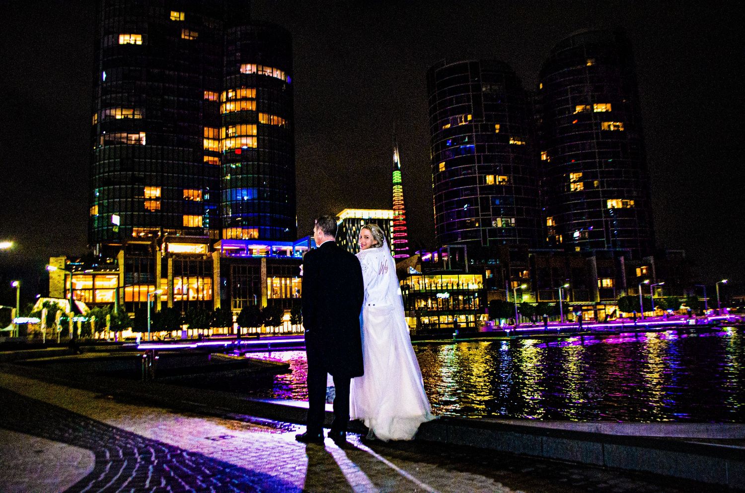 Couple in wedding attire poses against illuminated city skyline at night.