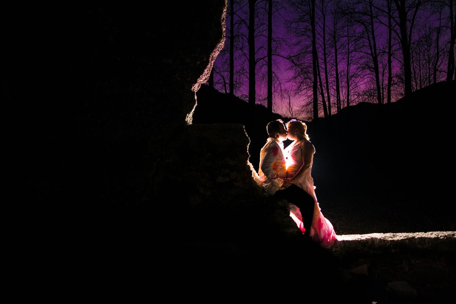 Backlit couple in dramatic pose against purple sky with silhouetted trees.