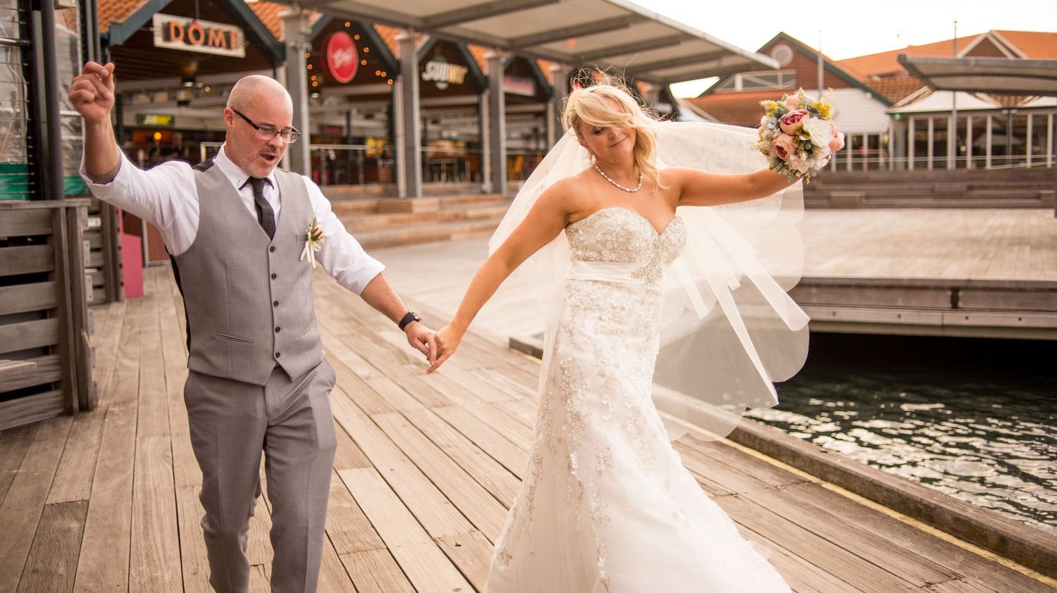 Wedding couple shares joyful moment on wooden boardwalk near waterfront shops.