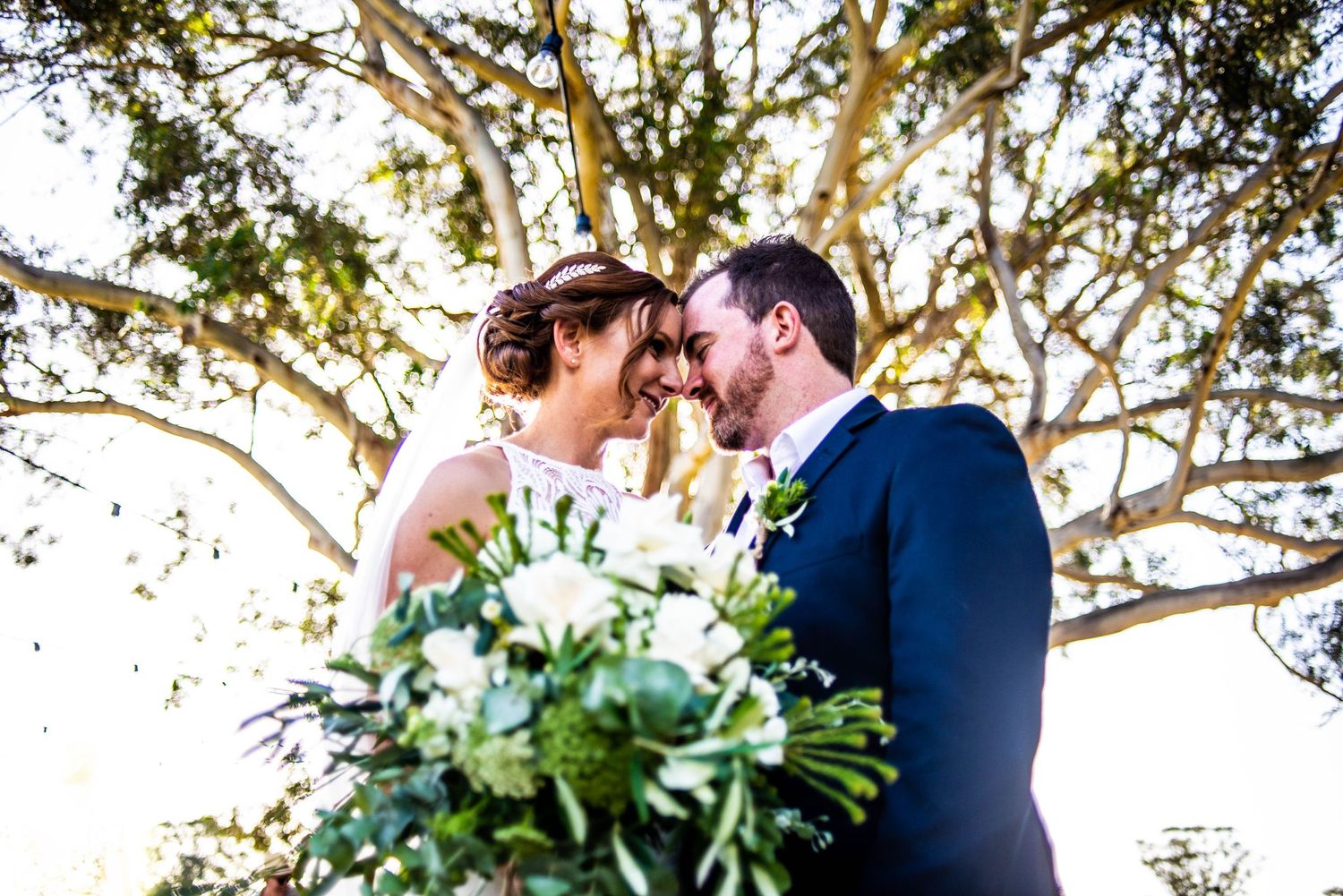 Wedding couple shares intimate moment under tree branches with white floral bouquet.