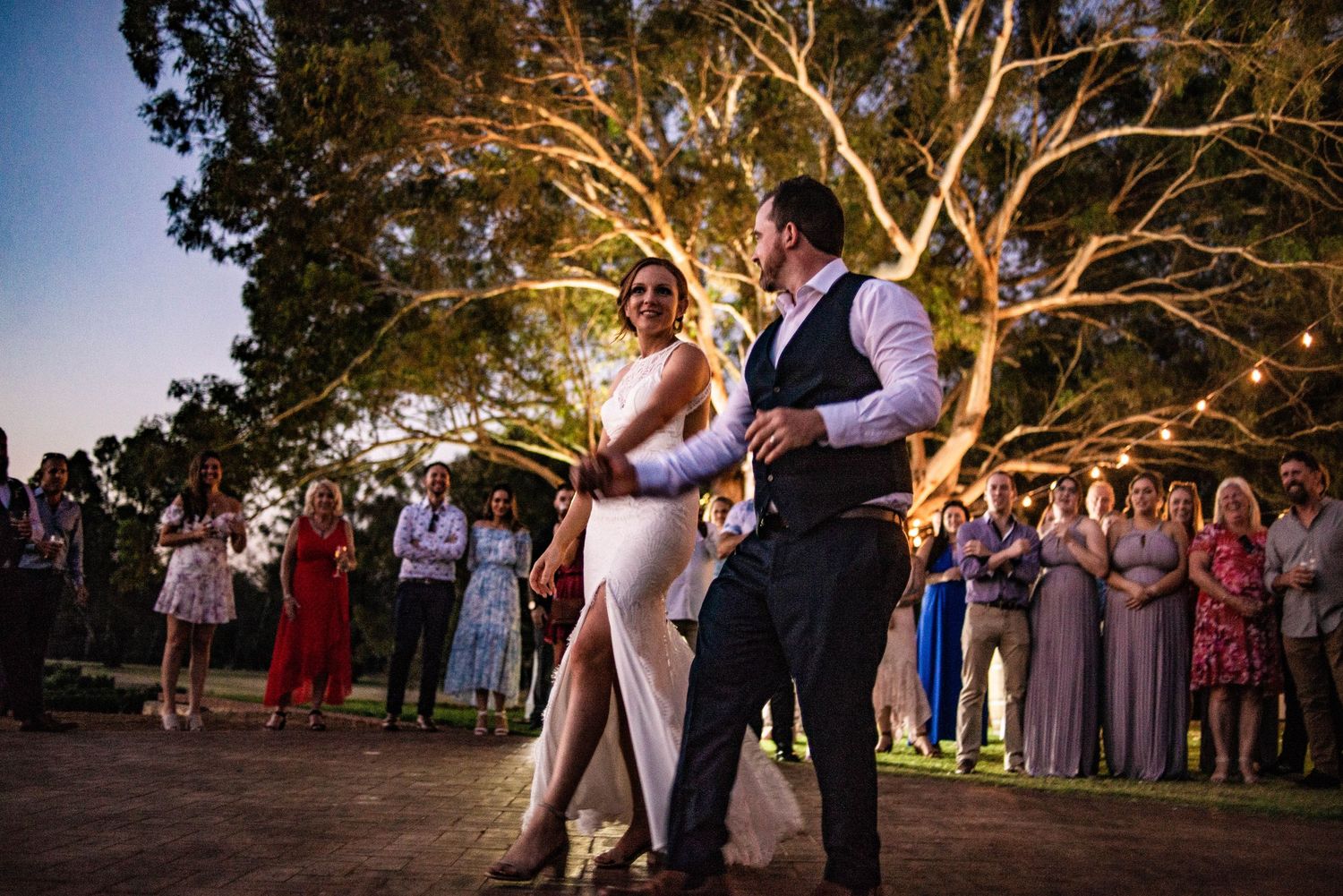 Wedding couple performs first dance outdoors at sunset with guests watching in background.