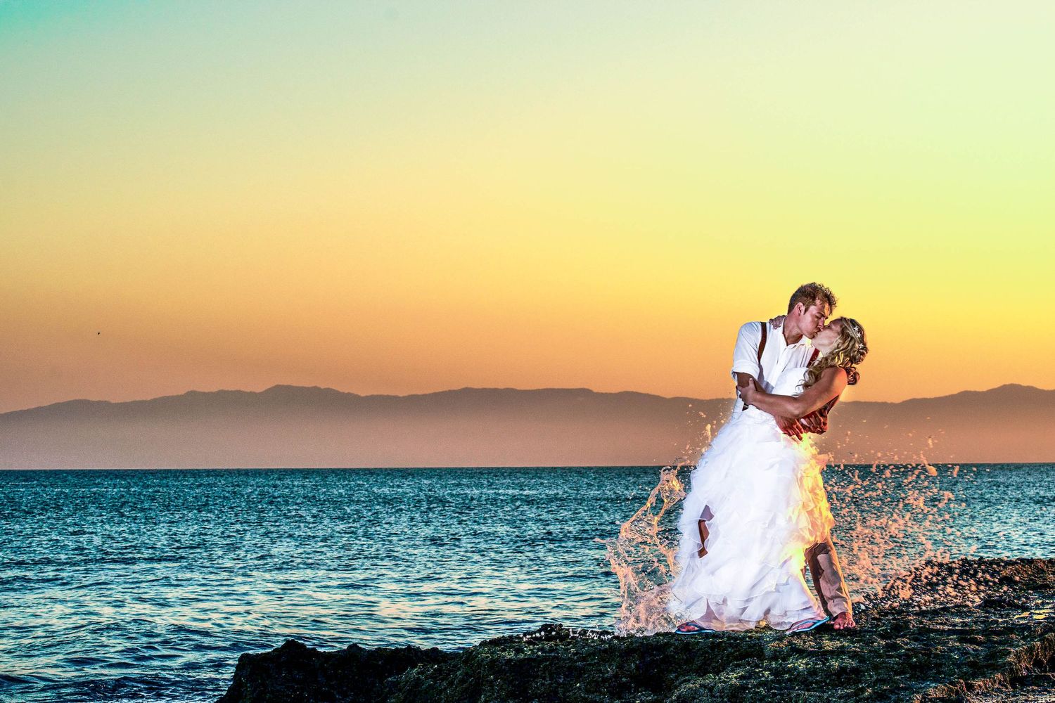 Couple in wedding attire embraces dramatically against colorful ocean sunset backdrop.