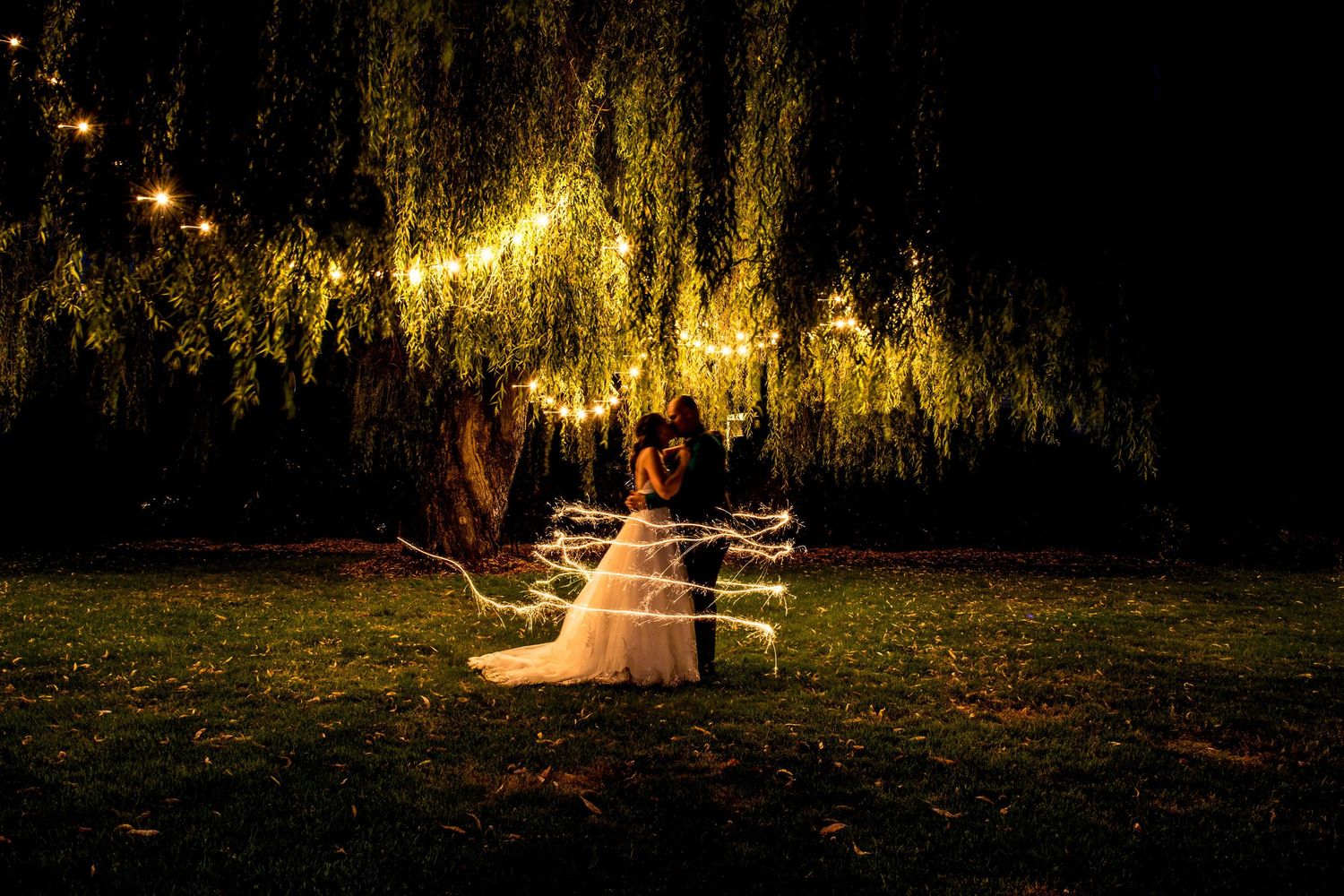 Romantic wedding couple sits on bench under illuminated willow trees at night creating magical ambiance.