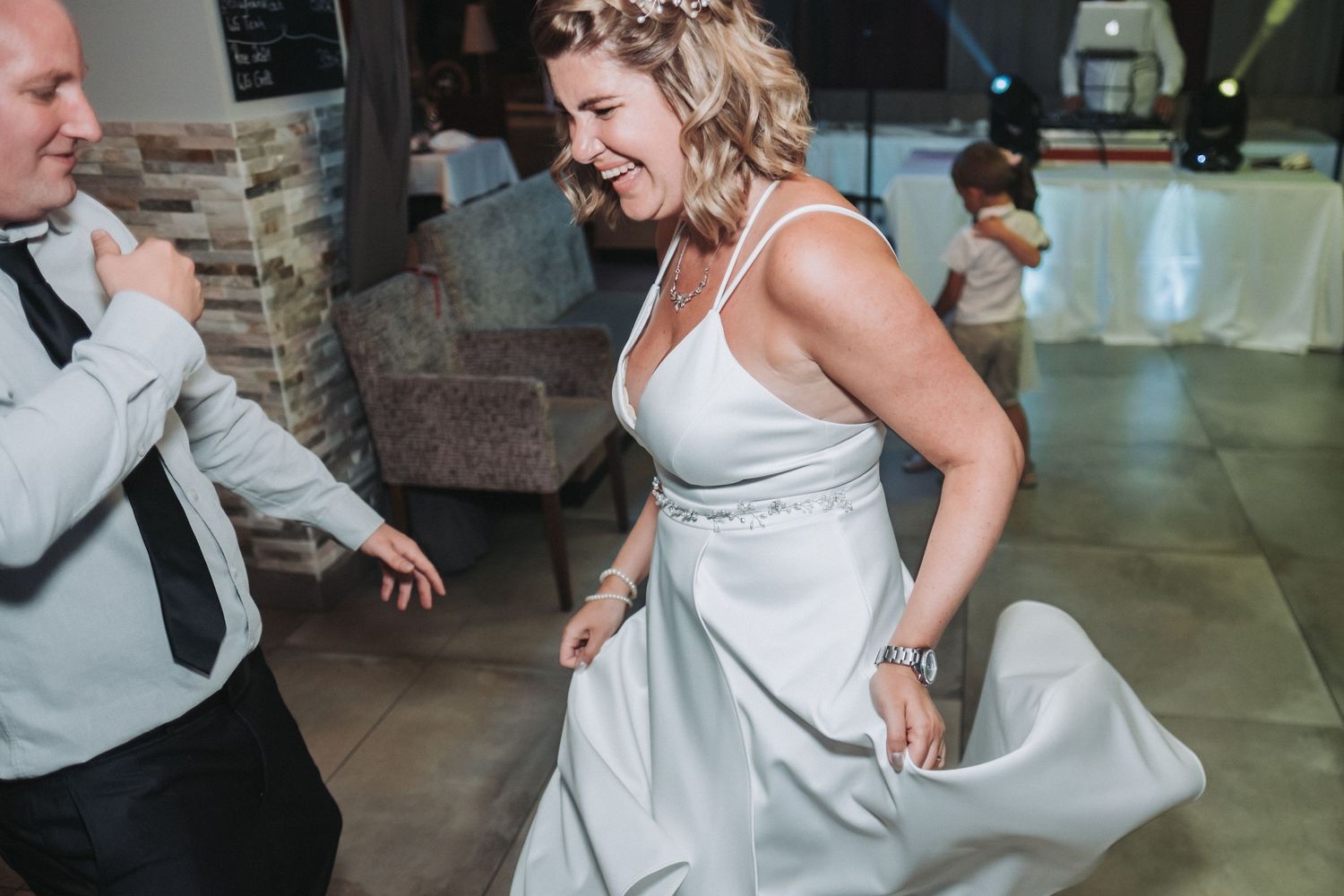 A bride in a sleeveless white dress dances energetically on the reception dance floor.