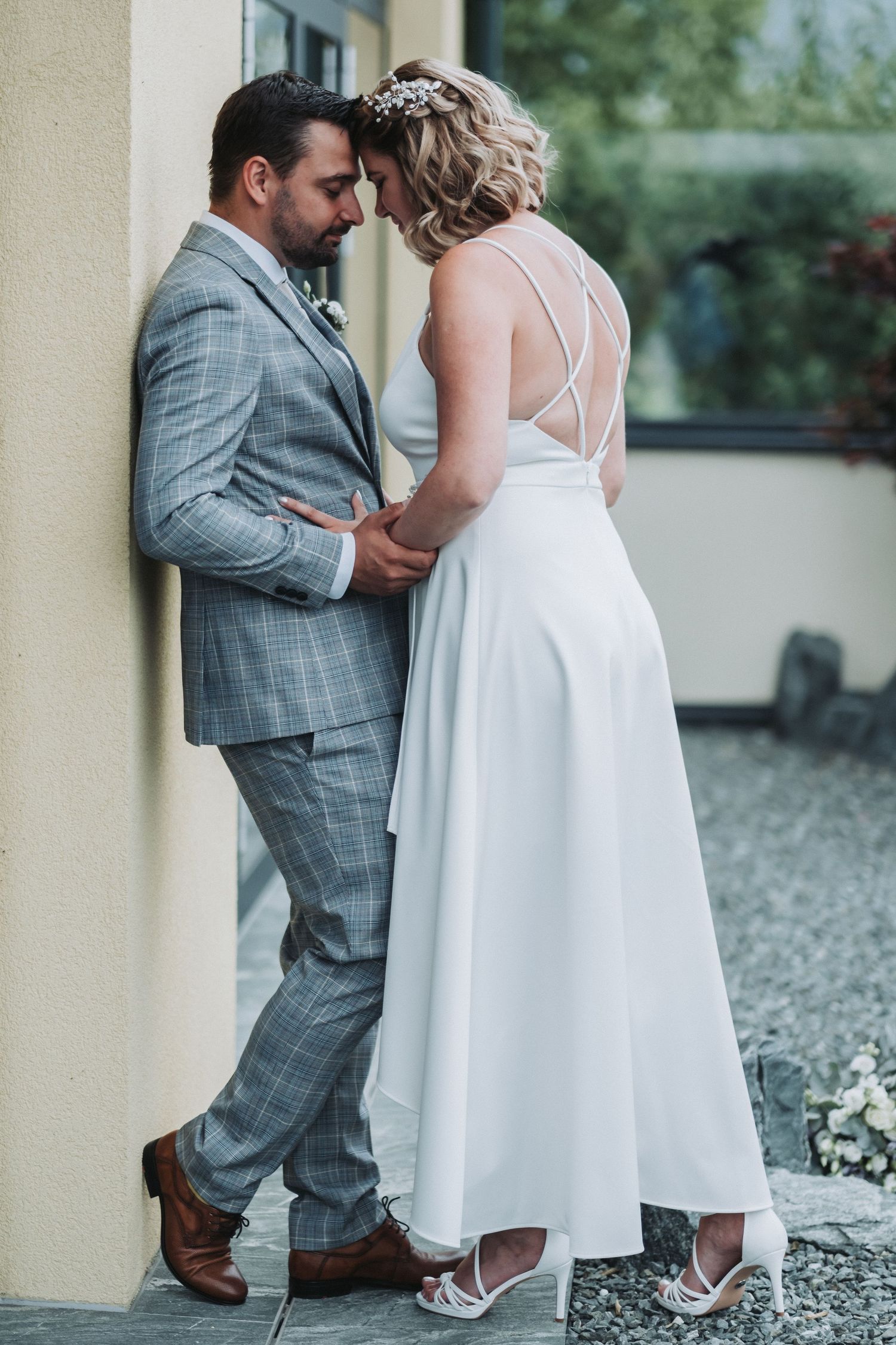 Tender moment between newlyweds in elegant attire against neutral wall backdrop.