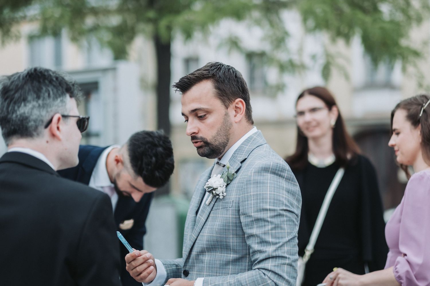 Wedding guests gathering outdoors with person in grey suit at center.