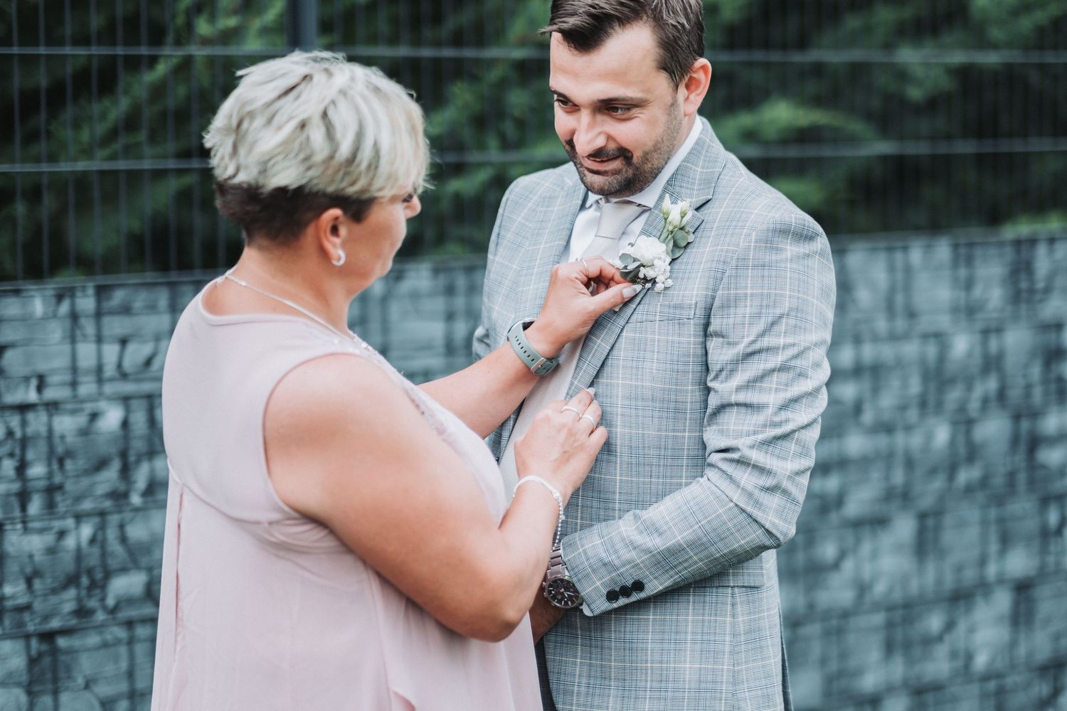 A boutonniere being pinned onto a light gray checkered suit jacket during a wedding preparation.