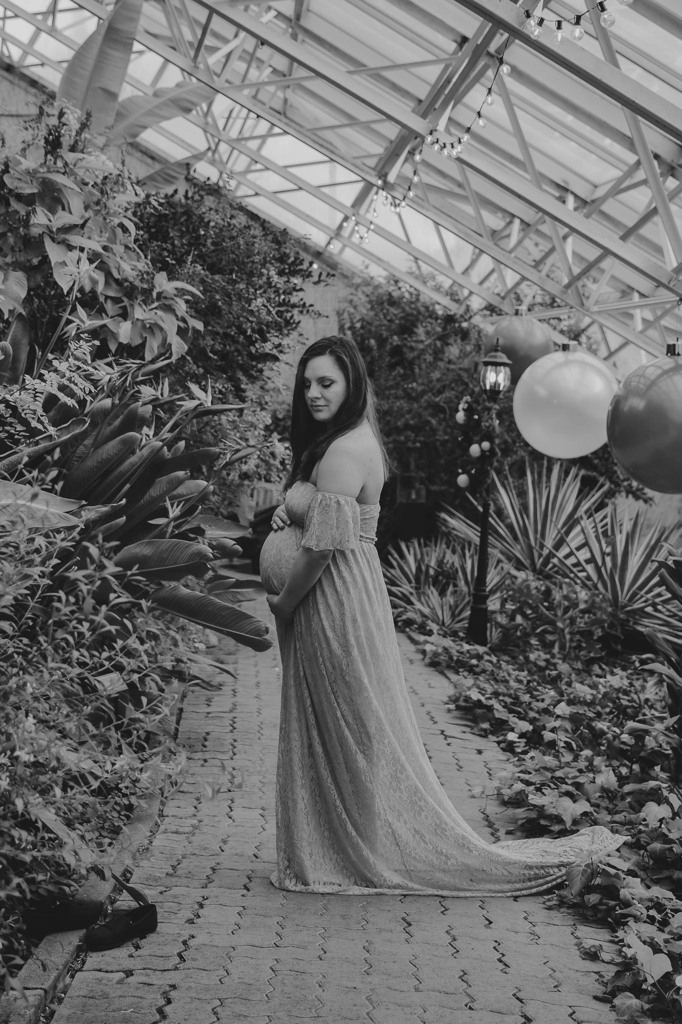 Black and white maternity photo showing an elegant dress in a greenhouse with balloons.