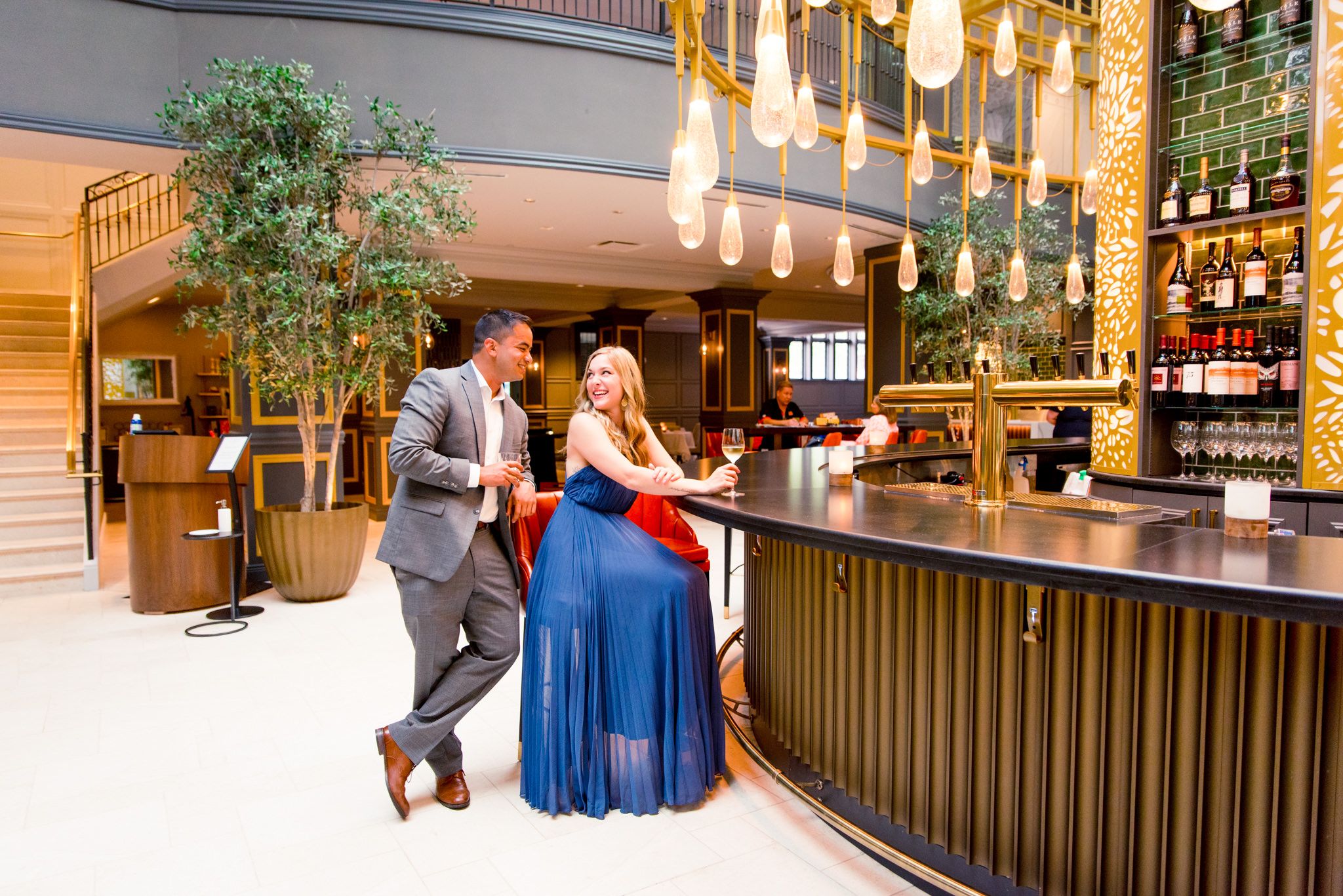 blonde woman in blue dress sits at the bar at the Lytle Park Hotel, laughing with fiance in grey suit standing behind