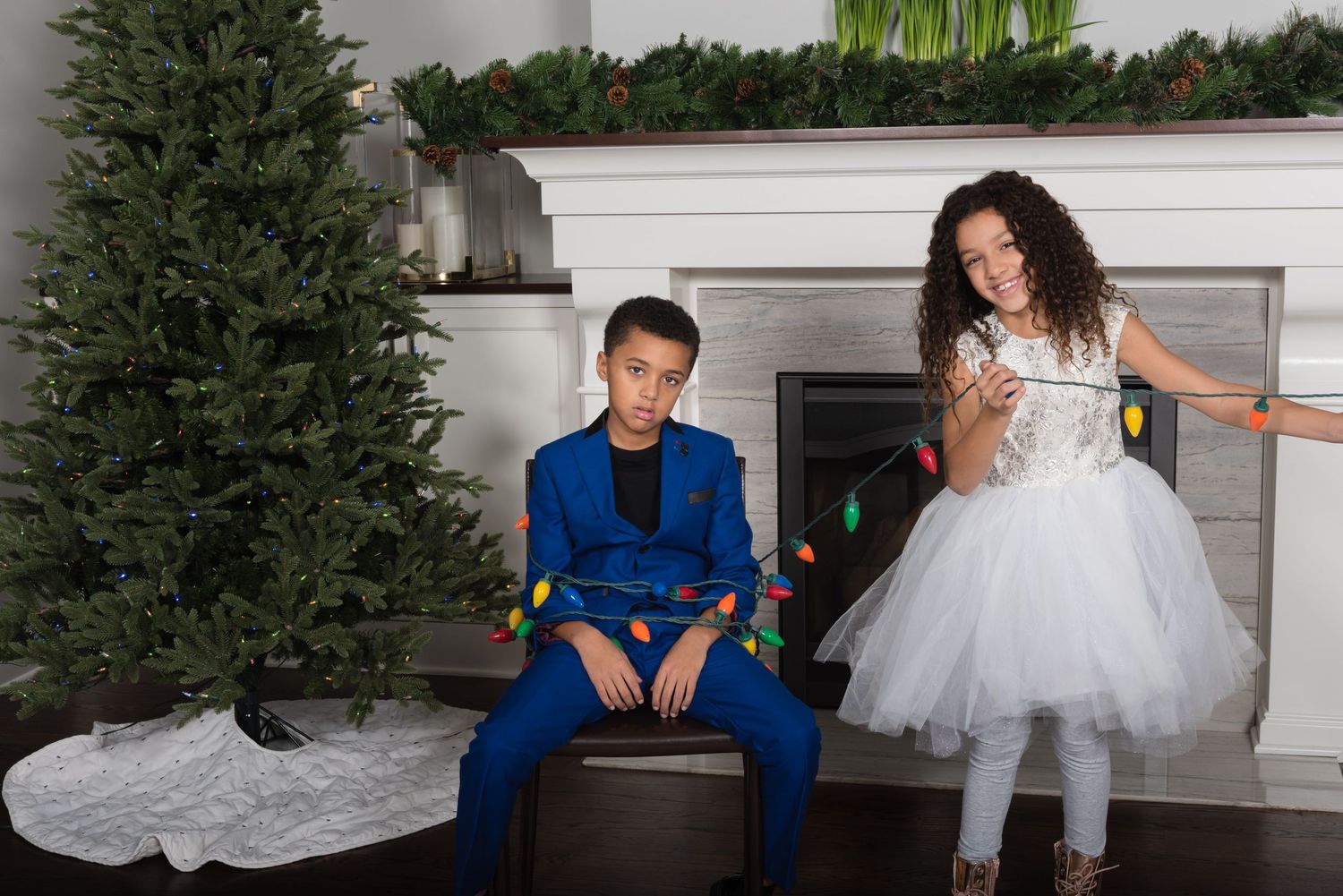 Two children in formal attire pose by Christmas tree and fireplace.