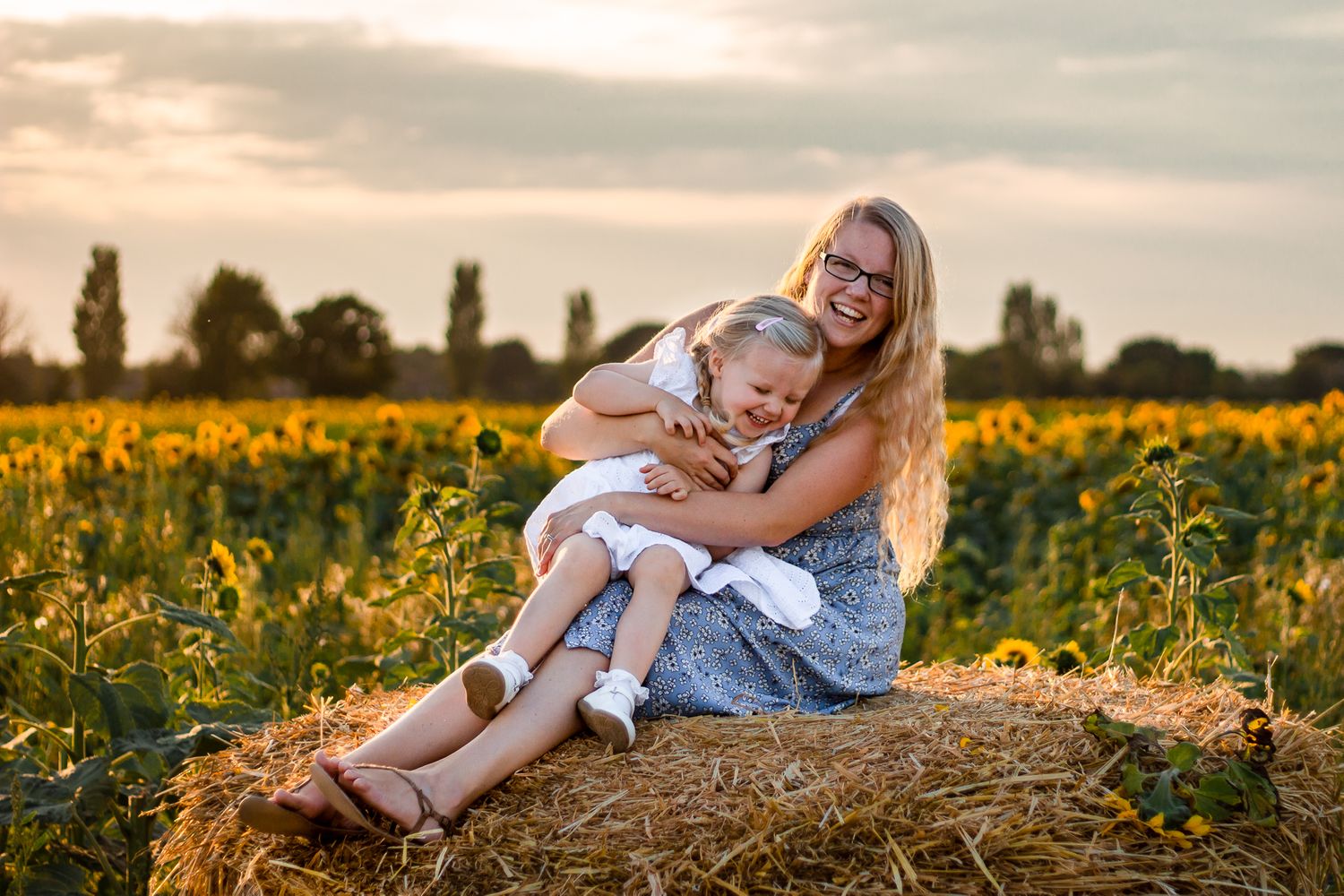 In a sunflower field on Hayling Island at sunset a Mum and her 2 year old daughter sit on a haybale laughing.