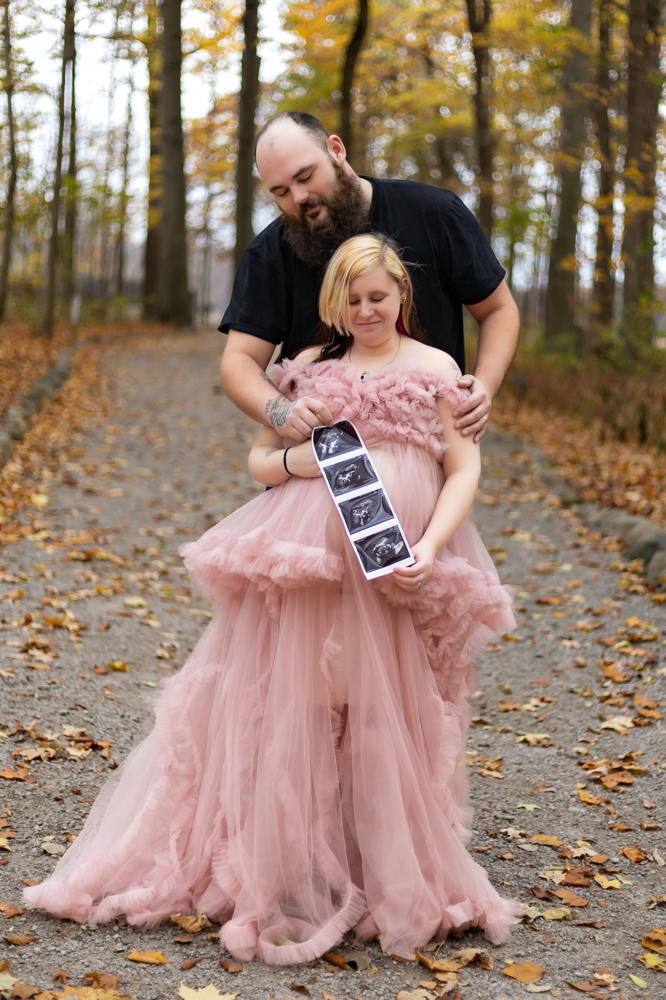 A couple poses with ultrasound photos in autumn foliage while wearing a flowing pink dress.