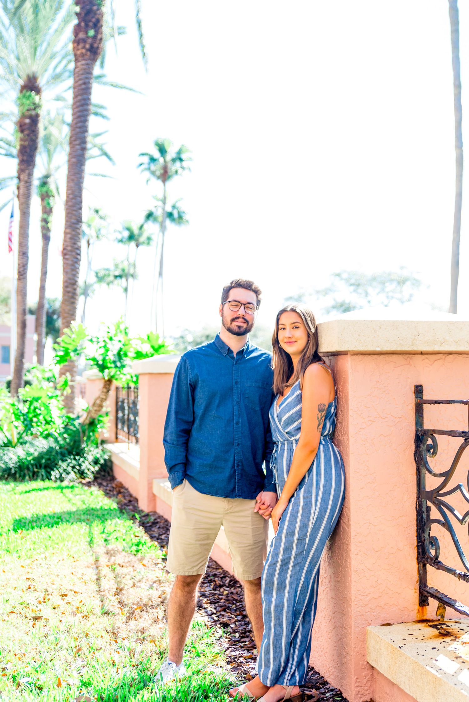 young woman and man hold hands against a pink wall under palm trees in Vinoy Park sunrise St. Pete engagement pictures