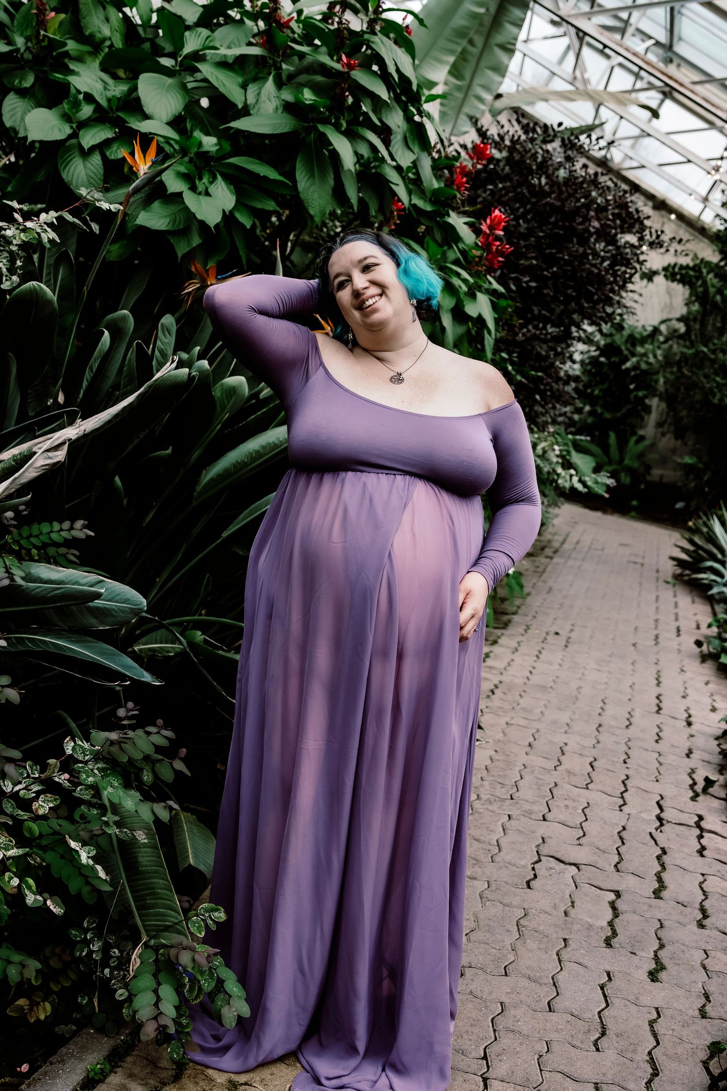 A purple off-shoulder maternity gown worn in a lush greenhouse setting with brick pathways.