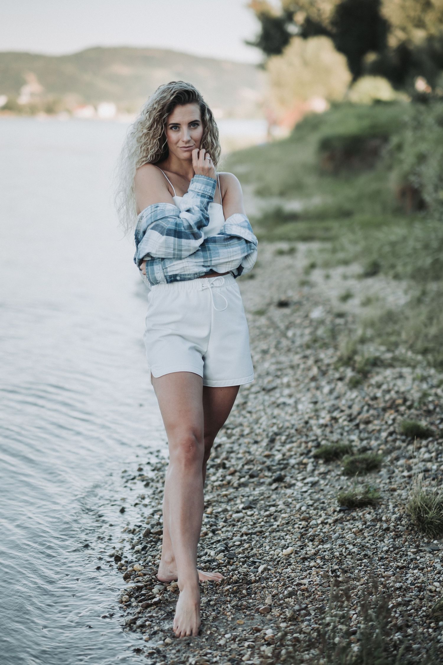 Woman in white shorts and plaid shirt poses barefoot by lakeside.