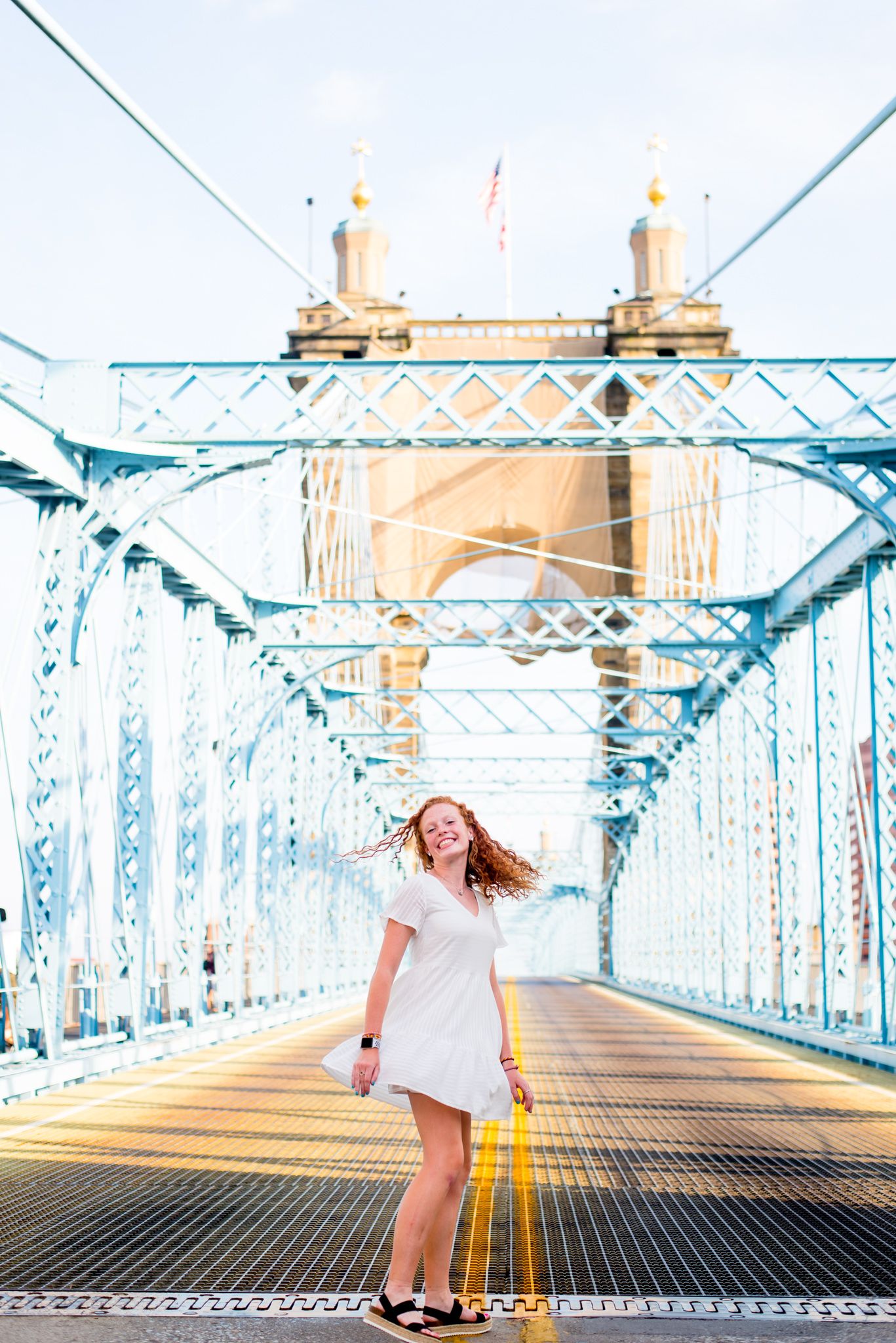 young woman with red curly hair in a white dress spinning around in the middle of Roebling Bridge for senior pictures